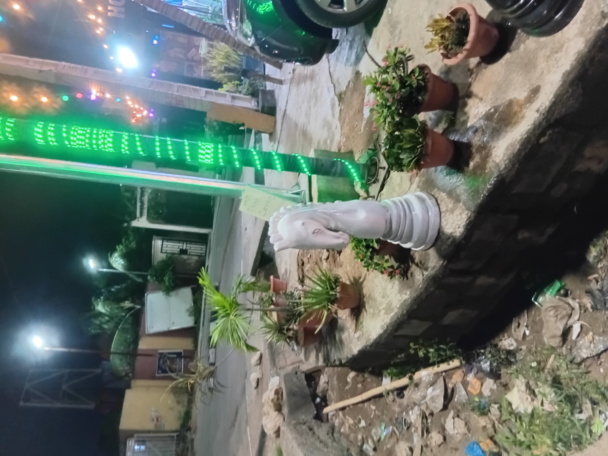 The image shows a nighttime scene in Ikeja, Nigeria. The foreground is dominated by a partially visible, large, white chess piece (knight) positioned on a ledge or step. Several potted plants are arranged nearby. The background includes a street illuminated by streetlights, various buildings, and some vegetation. A vehicle is parked partially in the frame at the top, and a structure with green lights is visible in the middle distance. The general condition of the immediate environment appears somewhat unkempt, with scattered debris and vegetation along the ledge and surrounding area. There are no people visible in the shot.