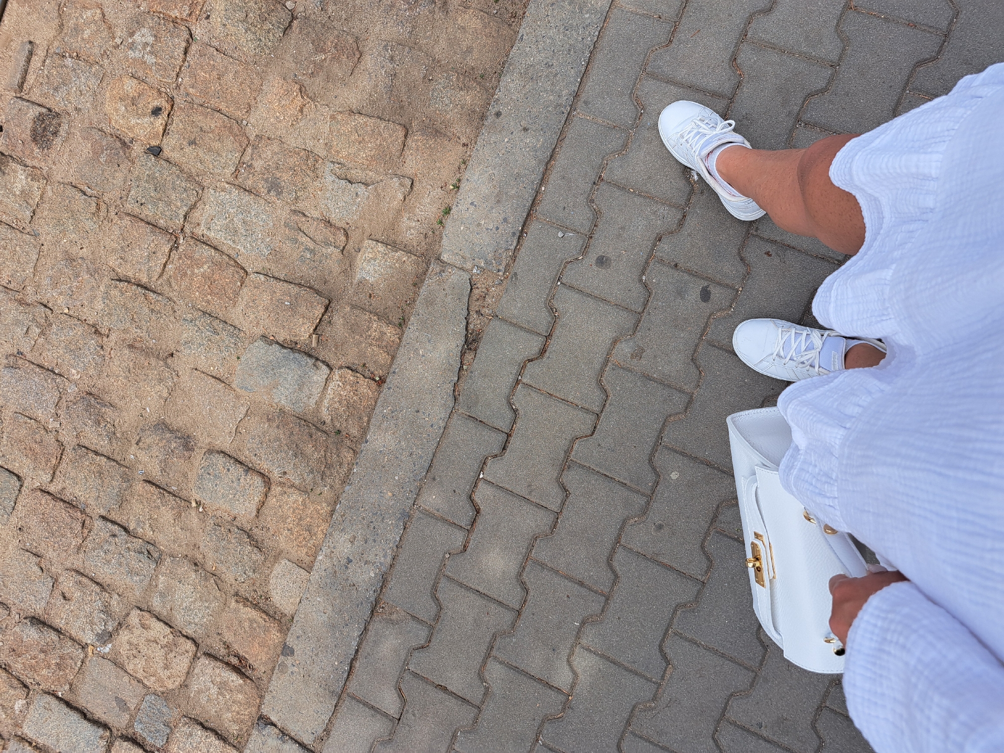 The media file is an overhead shot of a person walking on a cobblestone and paver sidewalk. The person is wearing white sneakers and a white dress, and is carrying a white handbag with gold hardware. The scene is outdoors, and the lighting suggests it is daytime. The ground is a mix of rough, tan cobblestones on the left and gray interlocking pavers on the right, separated by a concrete curb. The focus is on the person's legs and feet, as well as their handbag. The image is taken from a high angle, looking down. No text is visible in the image.