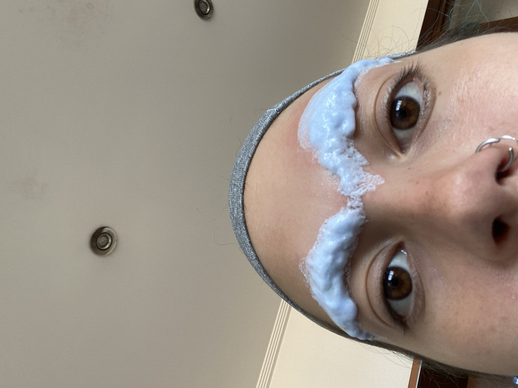 A close-up, tilted, selfie-style shot focuses on a person's face with a blue and white substance applied to their forehead, positioned above their eyebrows. The individual has brown eyes and a nose ring in their left nostril. They are wearing a gray headband. In the background, a plain, light-colored ceiling is visible with two recessed lighting fixtures. The location or any connection to Gijón/Xixón, Spain, is not evident within the visual information of this picture.