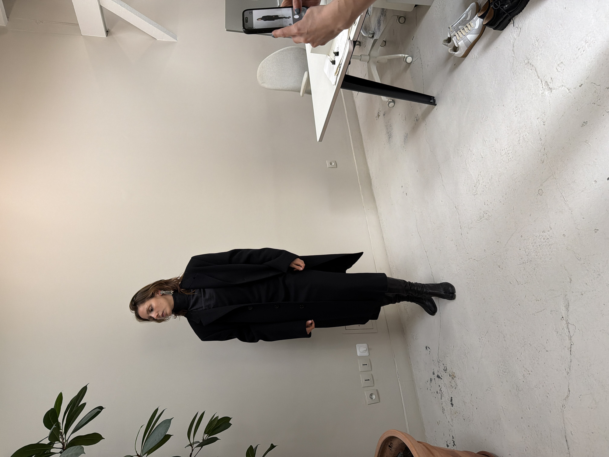 The image shows a woman in a black coat and boots standing against a wall in a room with a plant. Someone is taking a picture of her with a smartphone. The room contains a white desk with a chair and some shoes. The location might be Paris, France.