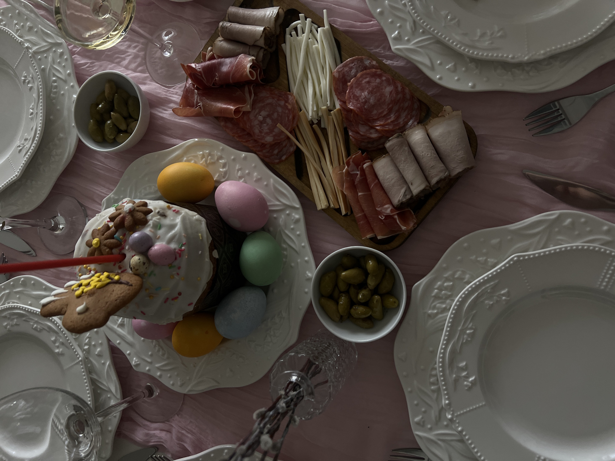A high-angle shot reveals a set table, likely for a festive meal, set against a light pink fabric backdrop. The table is populated with white, ornate plates arranged in layers. A central dish holds a cake decorated with white icing and multicolored sprinkles, surrounded by colored eggs in yellow, pink, green, and blue. A gingerbread cookie attached to a red straw rests near the cake. A wooden board contains an assortment of meats, including sliced salami, prosciutto, and rolled meats. Two small white bowls hold green olives. Glassware, including wine glasses and possibly other drinking vessels, are scattered across the table. Silverware is placed neatly next to the plates. The location appears to be indoors, and while Khimki, Russia, is specified, no specific details confirm this location. No people are visible in the frame.