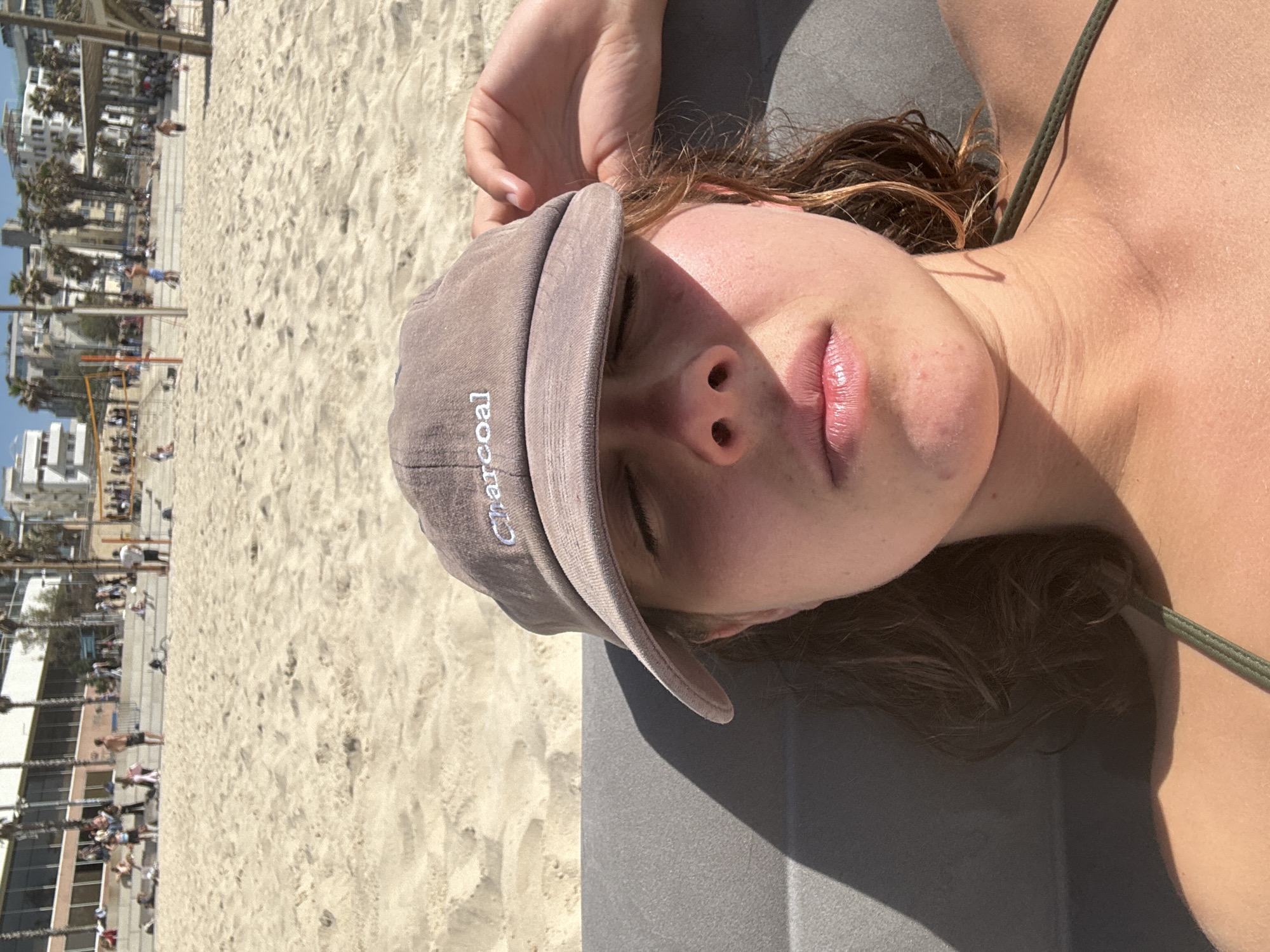 The image presents a close-up, first-person perspective of a young adult female resting on a beach, with a bustling urban beach scene in the background.
In the foreground, a female subject is seen from the chest up, reclining on a dark grey textured surface, possibly a beach chair. Her eyes are closed or squinting, with a taupe baseball cap bearing the white embroidered word "Charcoal" pulled low over her forehead, casting a shadow on the upper part of her face. Her lips are slightly parted and appear glossy. Her skin exhibits a light tan with some minor red blemishes on the chin area. Dark, possibly wet, hair frames her face. A dark green strap of a swimsuit or top is visible over her right shoulder/collarbone. Her right hand is partially visible, raised near her temple.
The midground and background reveal a wide, sandy beach under bright daylight. Numerous blurred figures of individuals are scattered across the light-colored sand and on an elevated, multi-tiered concrete or paved promenade that separates the beach from the city. Some people are standing, others are seated or walking. A volleyball net is visible on the sand to the right. Behind the promenade, multiple modern, multi-story buildings with balconies line the beachfront, interspersed with numerous tall palm trees. The sky above is clear and blue.
The scene depicts a public beach in the city of Tel-Aviv, Israel, identifiable by its urban beachfront architecture, characteristic palm trees, and public recreational facilities.