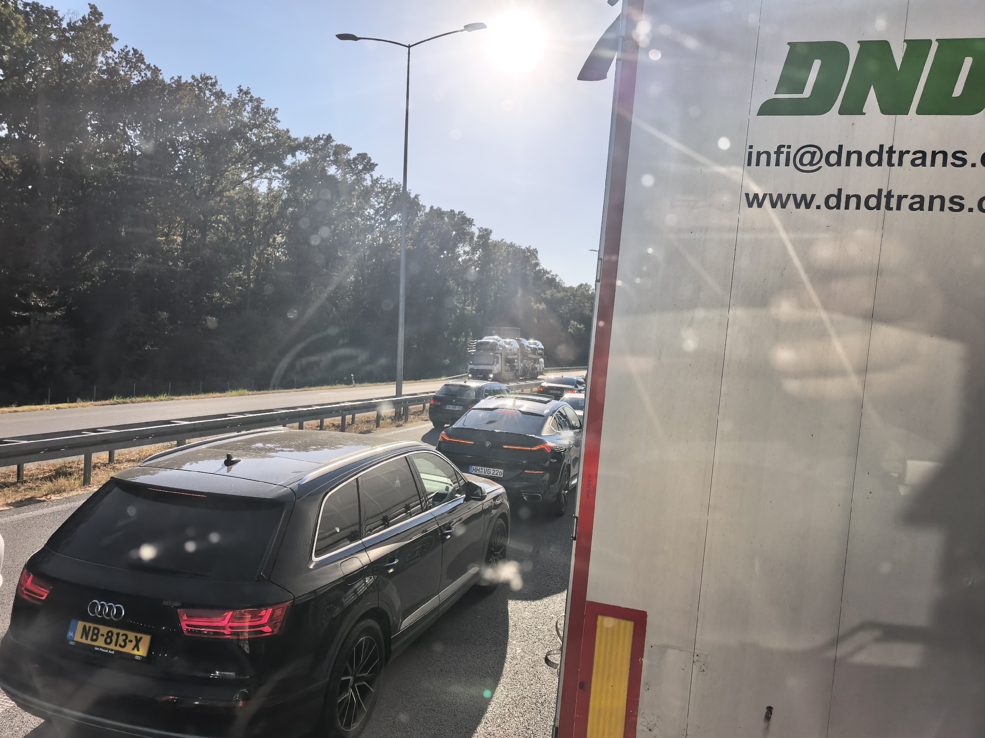 The image shows a traffic jam on a sunny day. A black Audi Q7 is in the foreground, with its rear license plate reading "NB-813-X". Behind it, several other cars are visible, including a black BMW X6 with license plate "HH:VG 226". Further ahead, a car carrier truck is transporting other vehicles. The scene appears to be on a highway, with trees lining the sides and a bright sun in the sky. The right side of the image shows the side of a white truck with the green "DND" logo and website "www.dndtrans.com". Based on the license plates and the overall environment, the location is likely a highway in Europe, possibly within or near Sid Municipality, Serbia, given the provided context. However, the image itself does not contain specific landmarks to confirm the exact municipality.