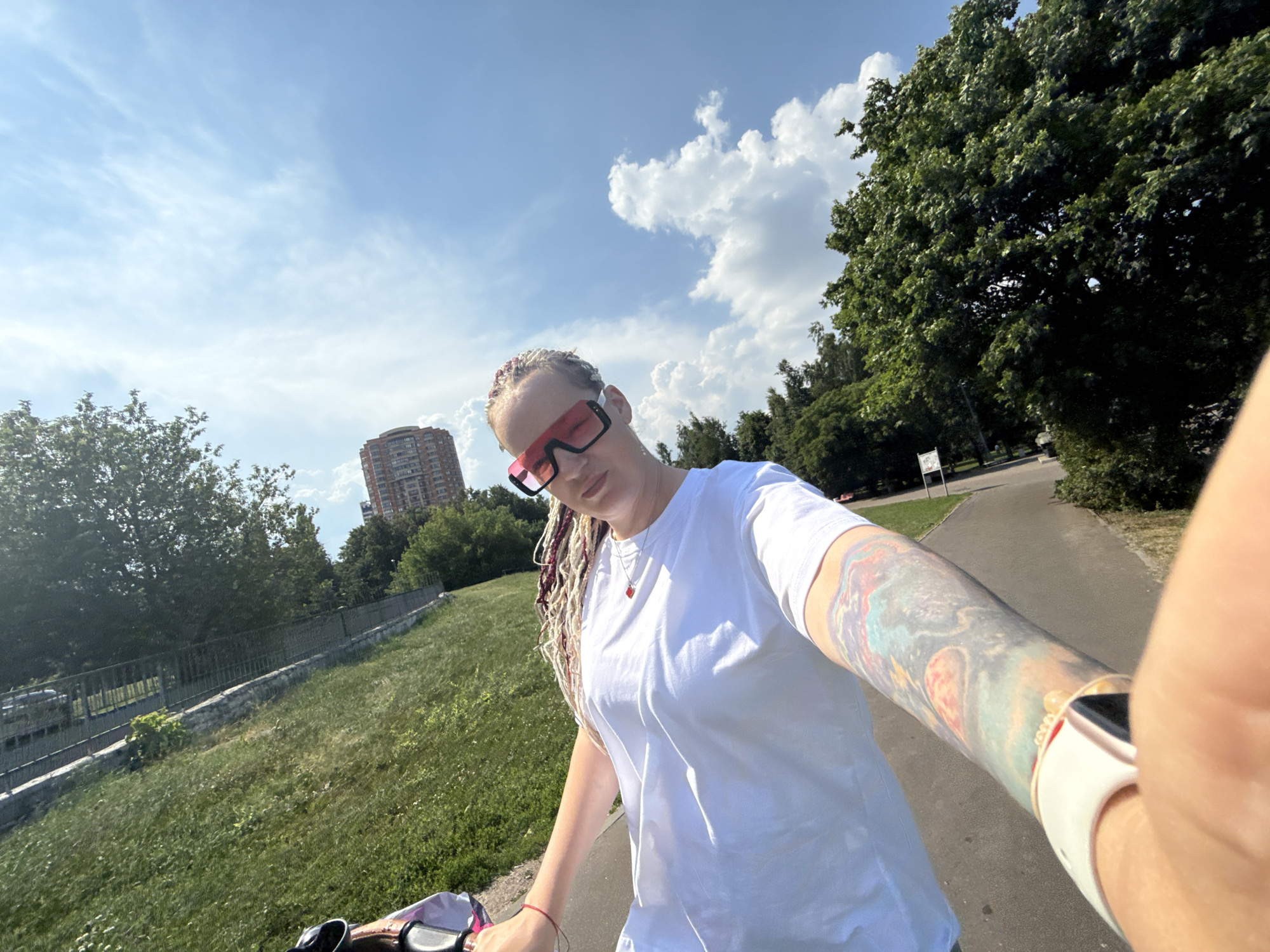 A selfie taken from a low angle shows a person with blonde braids, some dyed pink and red, wearing oversized pink-framed sunglasses and a white t-shirt. The person's left arm is heavily tattooed with a colorful cosmic design, and a white smartwatch is visible on their wrist. They are holding onto bicycle handlebars. The background features a grassy slope, a paved path, a metal fence, and lush green trees. In the distance, a tall, reddish-brown apartment building is visible against a bright blue sky with scattered white clouds. The scene appears to be in a park or a green space within a city. Given the architectural style of the building and the general urban park setting, this could be a location in Moscow, Russia, such as Ostankino Park or Sokolniki Park, which are known for their large green areas and tall residential buildings.