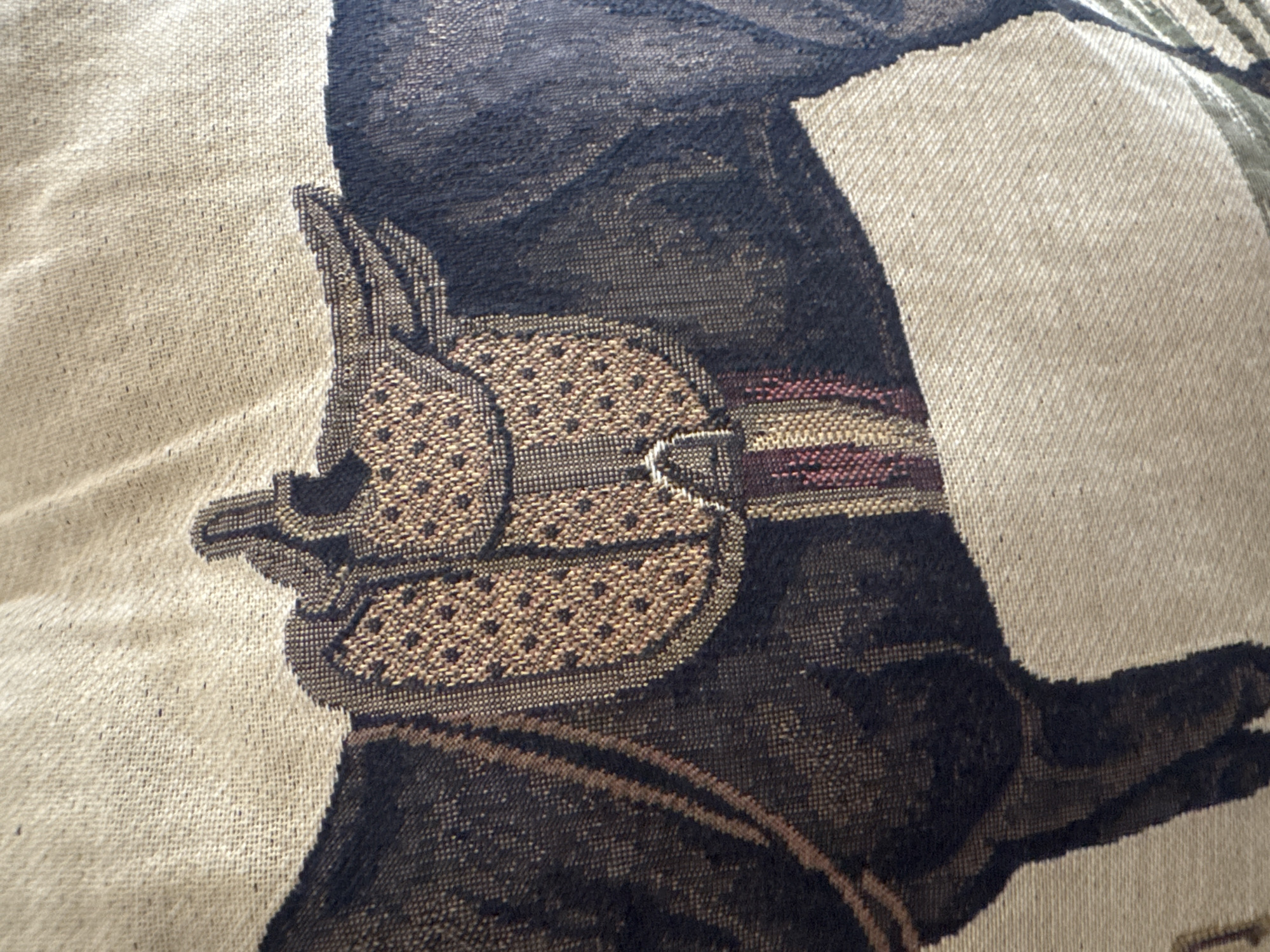 This media file is a close-up photograph of a woven textile, likely a tapestry, blanket, or cushion cover, showcasing an intricate design. The primary subject depicted is a stylized figure, seen in profile, facing towards the left. The figure appears to be a person, possibly a warrior or a historical individual, adorned with an elaborate headpiece that resembles a helmet or decorative headgear. The headpiece features detailed, repetitive patterns of small dots in light brown and gold tones, contrasting with the darker brown and black hues of the main figure. Straps with maroon and lighter brown stripes are visible across what appears to be the chin or neck area, connecting to the headgear.
The figure's upper body or shoulder area is also visible, woven in dark brown and black threads. The background of the textile is a plain, light beige woven fabric, providing a clear visual separation from the darker figure. The overall texture of the textile is prominent, highlighting the individual threads and the detailed weaving technique used to create the artwork.
There is no specific activity or event taking place; the image is a static artistic representation. The style suggests a cultural or historical motif. No visible text is present. Given the location context of Puebla, Mexico, the artwork likely draws inspiration from local cultural or historical narratives, possibly reflecting indigenous or colonial art forms.