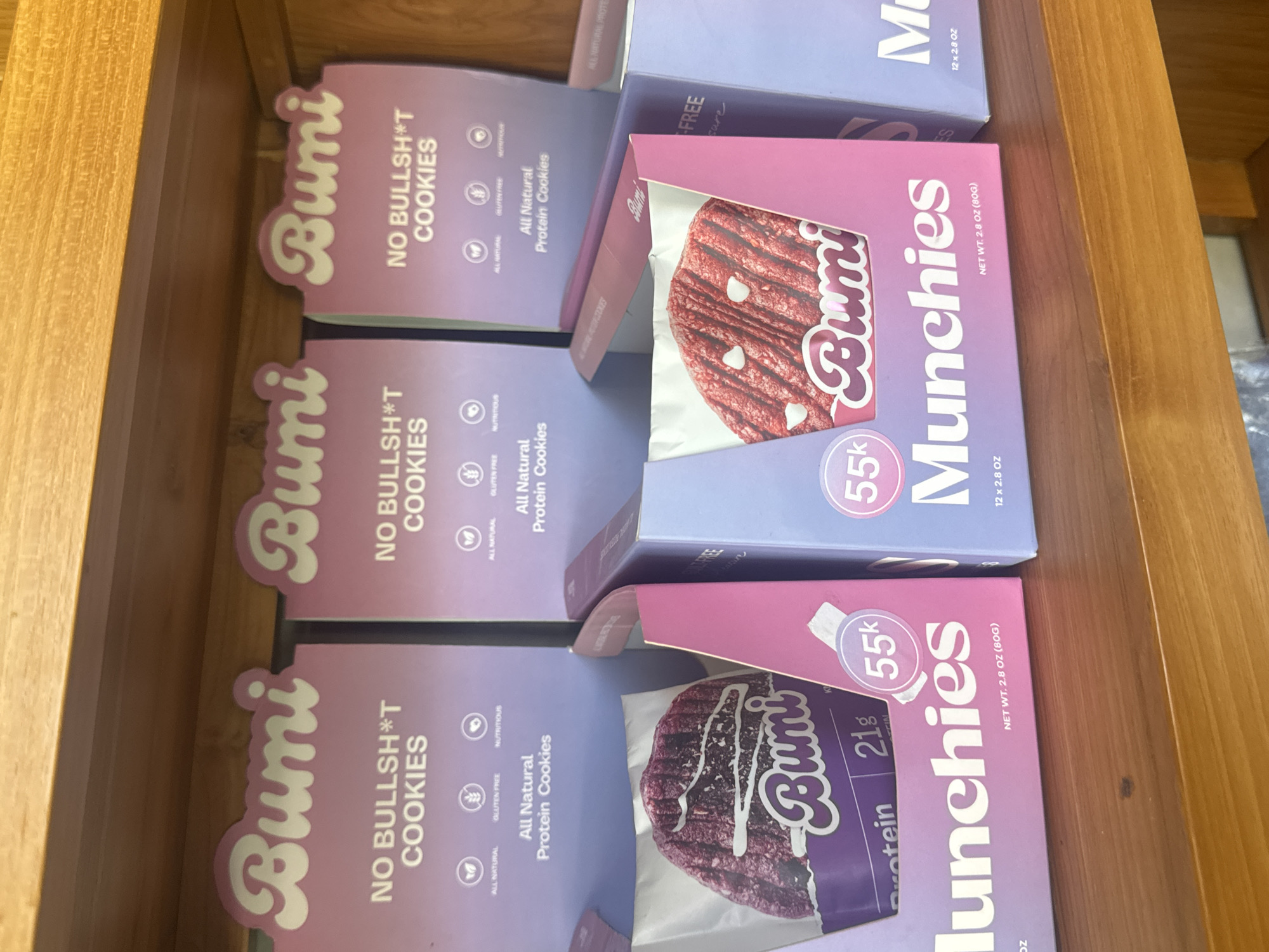 This image shows a drawer filled with boxes of "Bumi" brand cookies. There are two types of cookies: "No Bullsh*t Cookies" and "Munchies." The "No Bullsh*t Cookies" boxes are purple with the text "NO BULLSH*T COOKIES" and list that they are all natural, gluten-free, nutritious, and all-natural protein cookies. The "Munchies" boxes are pink and show images of the cookies. They list the weight of the contents as 2.8 oz (80G) and contain 21g of protein. The drawer is made of wood. It's impossible to determine the location in the city of Ubud District, Indonesia based on this image alone.