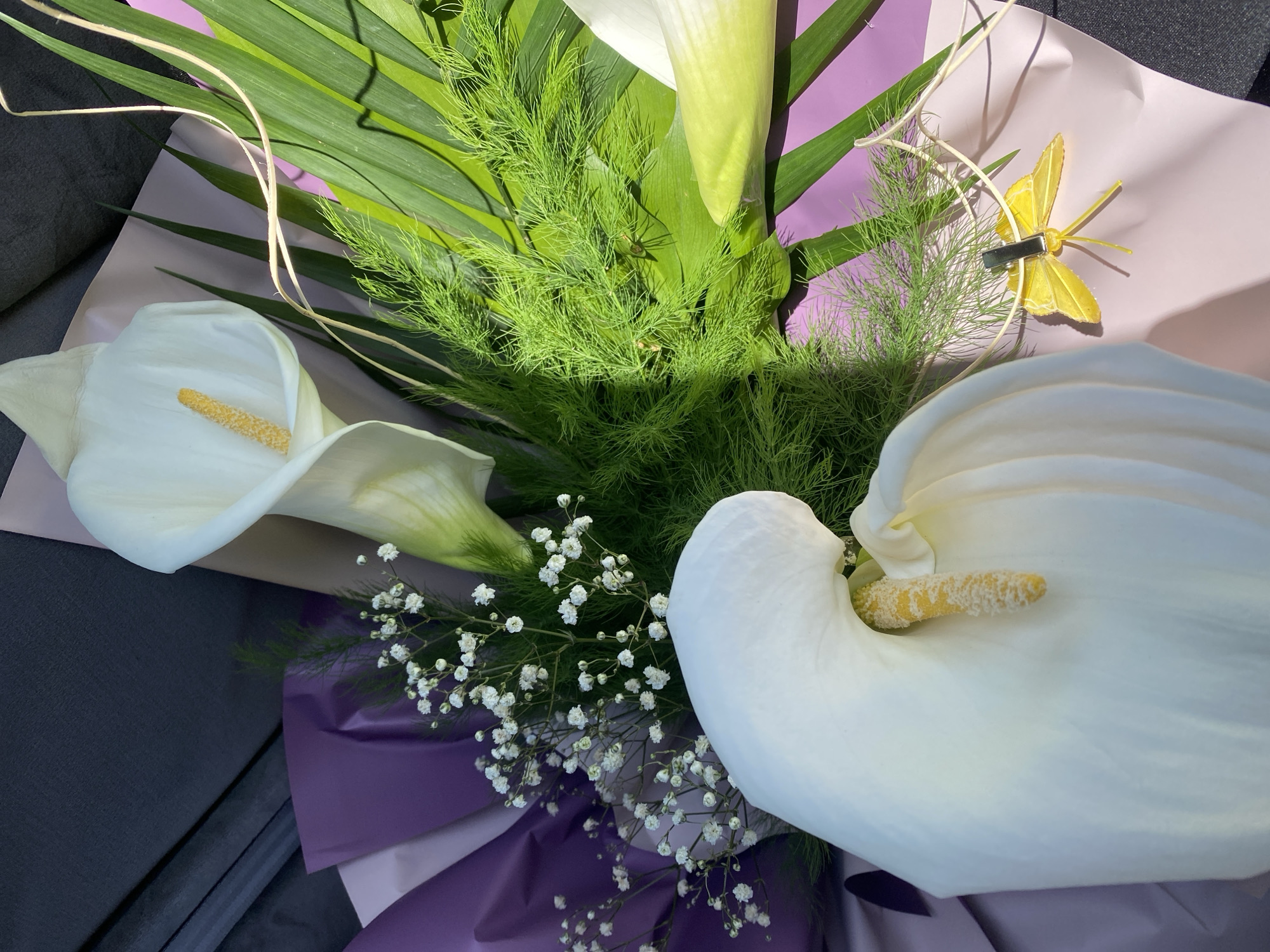 A close-up shot of a bouquet of white calla lilies and other greenery, wrapped in light pink and purple paper. A yellow butterfly decoration is attached to the bouquet. The location is unknown.