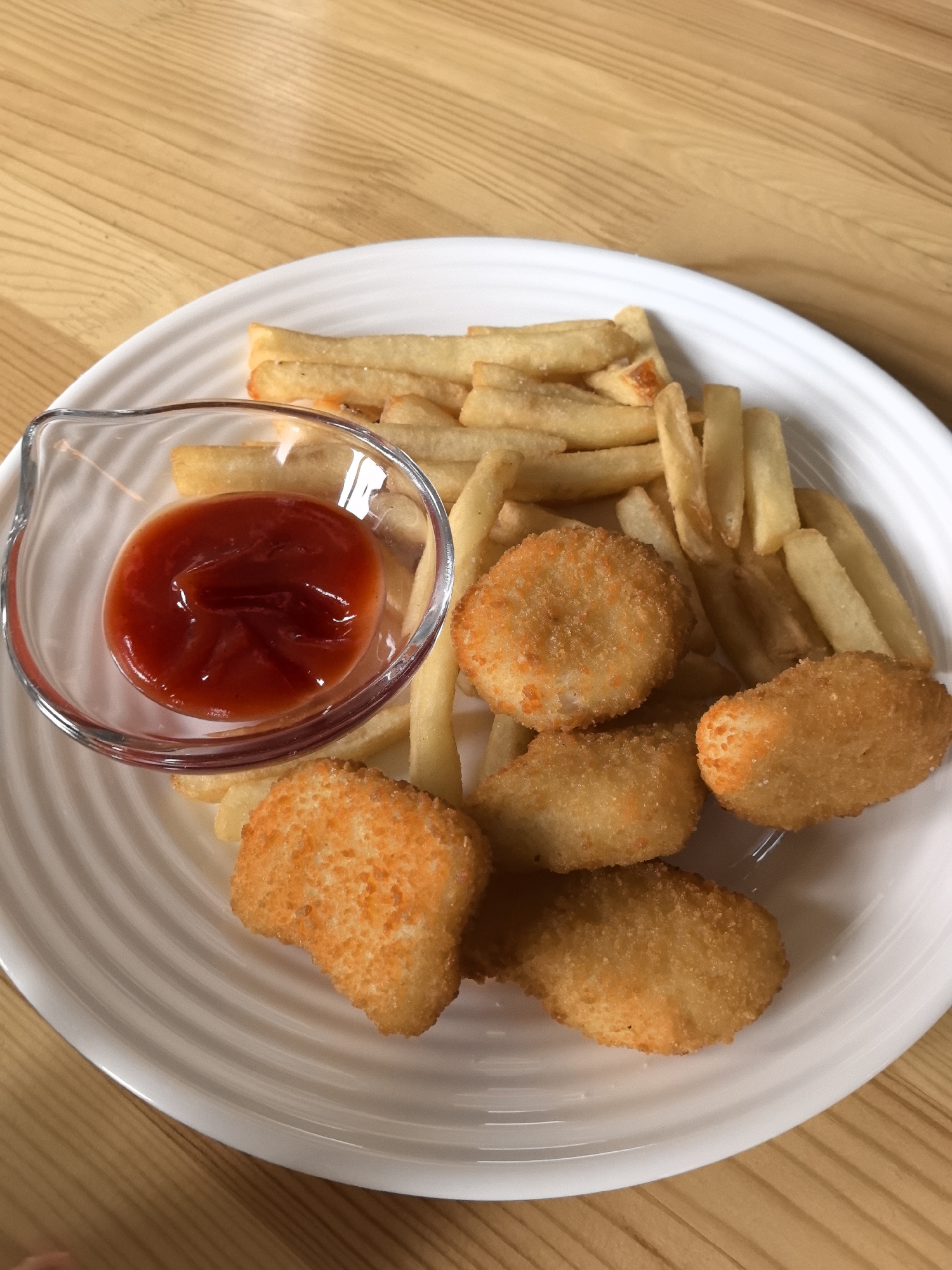 A white plate sits on a wooden table, containing a serving of french fries, chicken nuggets, and a small clear glass bowl of ketchup. The food appears to be on a table in Perm, Russia.