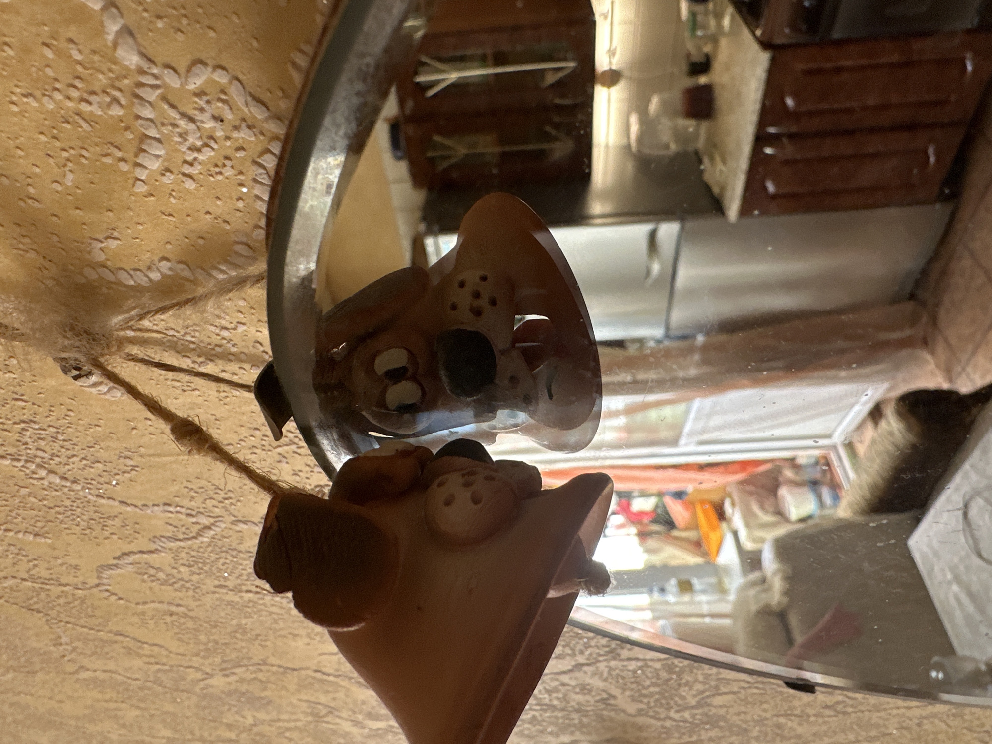 A terra cotta figure of an animal, possibly a fox or dog, is positioned near a circular mirror. The figure is detailed with pronounced eyes and a patterned snout. The mirror reflects the same figure, partially obscuring the view of objects behind it, including a set of wooden cabinets, various household items, and light filtering through a window. The background beyond the reflection gives a partial glimpse of a domestic setting. The surface on which the mirror and figure rest has a textured, possibly painted finish.