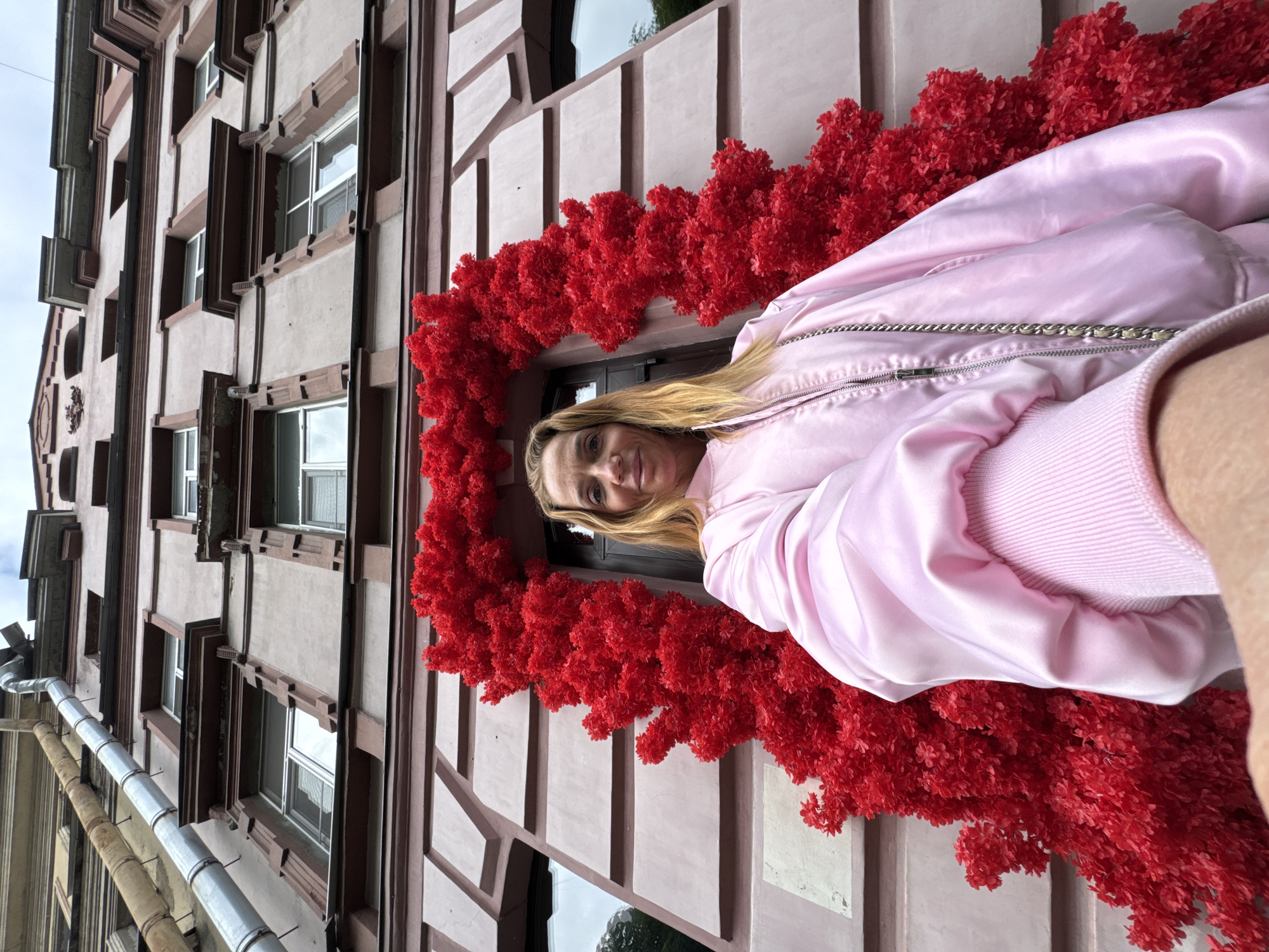 A medium shot shows a woman wearing a pink jacket in front of a window framed by a red floral arrangement. The window is set in a light beige building with dark brown architectural trim. Windows are stacked vertically and the building appears to be three or four stories tall. A metal pipe runs vertically along the left edge of the building. The image is angled, giving the impression of looking upward. The location is Saint Petersburg, Russia.