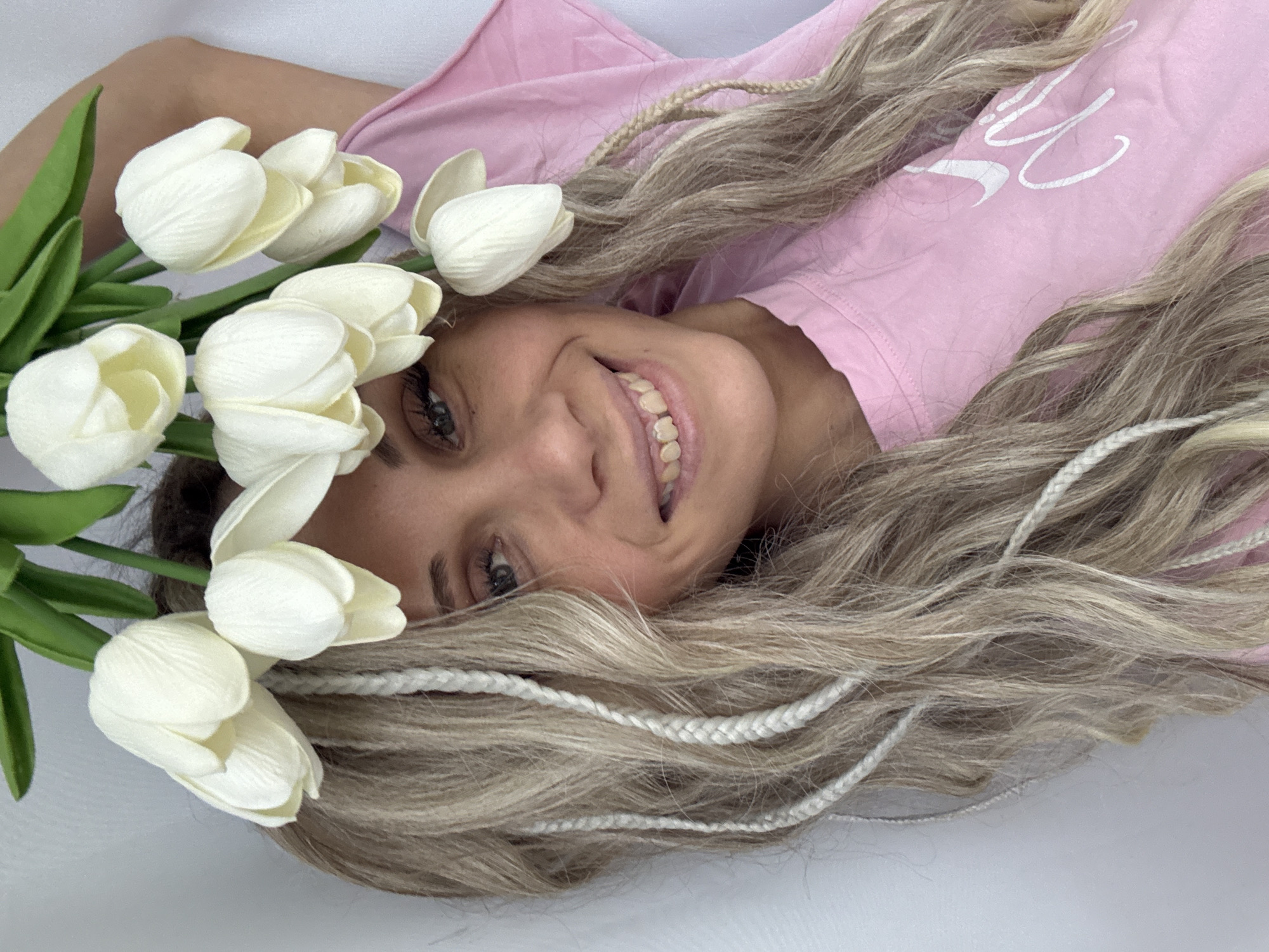 The image shows a smiling woman with blonde hair, braided with white threads, wearing a pink t-shirt. She is lying down, and a bouquet of white tulips is positioned near her head. The background is a plain white surface. No information about the city of Rusanovo, Russia, can be determined from the image.