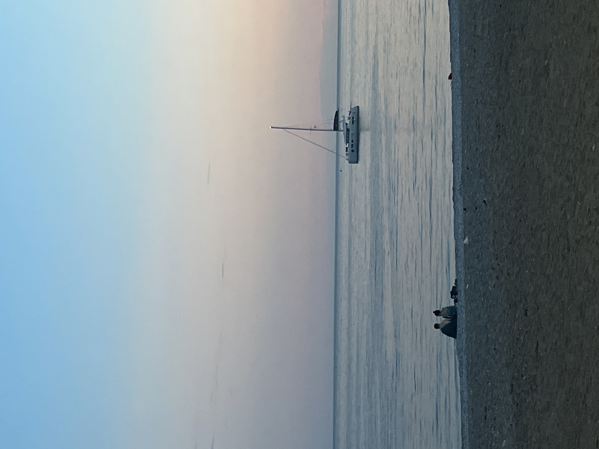 The image shows a sailboat in the sea off the coast of Budva, Montenegro. The sky is a soft gradient of blue, pink, and peach, suggesting either sunrise or sunset. The water is relatively calm, with subtle ripples. To the right of the frame is a concrete barrier or embankment with a few people seated on it.