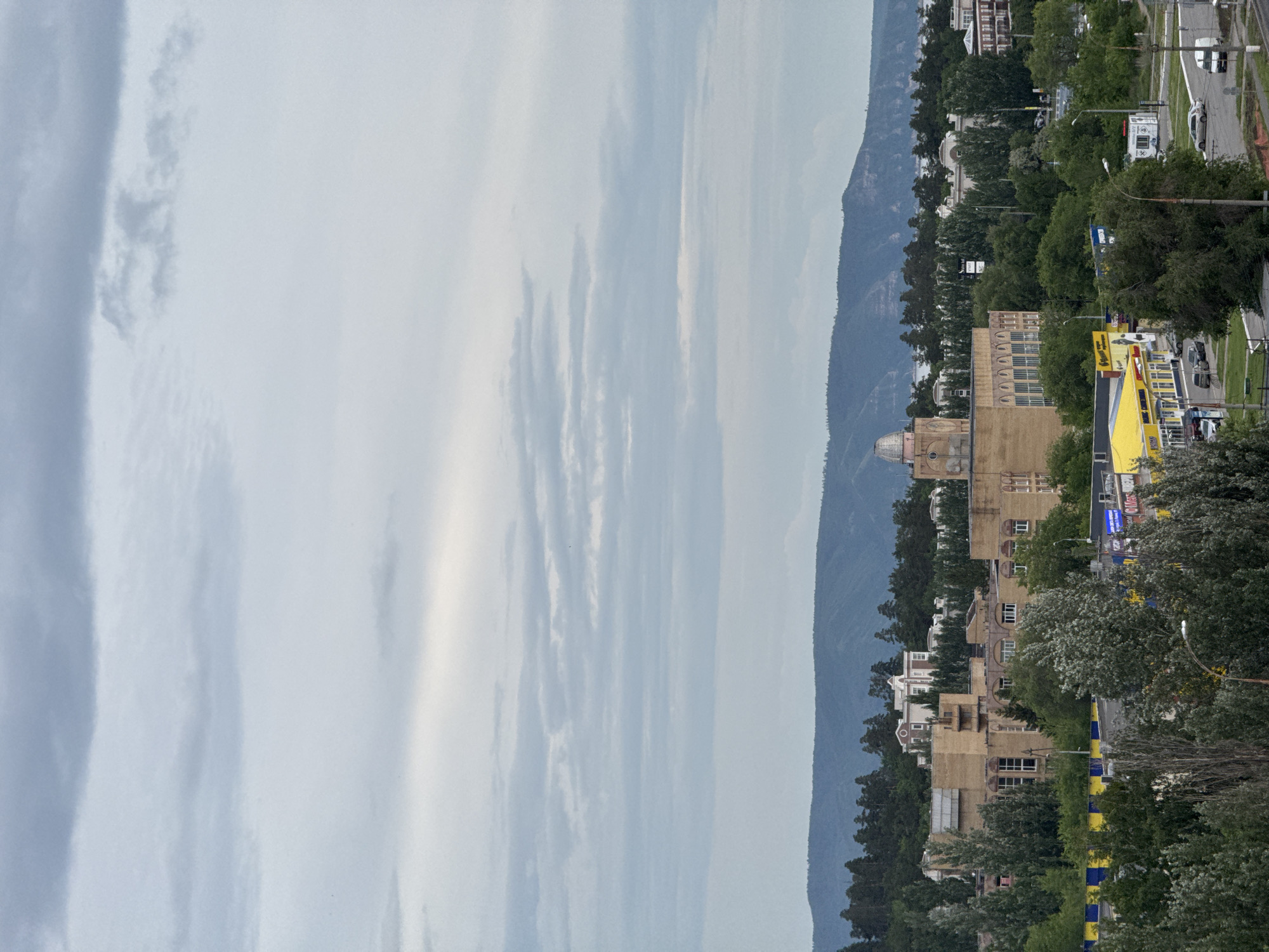 This wide-angle, high-elevation photograph captures an urban landscape under an overcast sky, with a mountainous backdrop. The scene is illuminated by diffuse daylight, indicative of a cloudy day with no direct sunlight.
The upper approximately 55-60% of the image is dominated by a pale gray, uniformly clouded sky, showing multiple horizontal layers of stratiform clouds.
In the background, a continuous range of dark, forested mountains or large hills stretches across the entire horizon. Below these mountains, a faint sliver of a body of water is visible, appearing lighter and reflecting the sky.
The midground features an array of buildings interspersed with abundant greenery. Prominently, a multi-story building with a light brown facade and a distinctive dome on its roof is located slightly left of the center. To its left, another large, multi-story building with a similar color palette and rectangular windows stands. Further left, partially obscured by trees, another multi-story building with a white, classical architectural style is visible on a higher elevation. Throughout this section, numerous trees, mostly deciduous, are interspersed, creating green patches and lining roads.
The foreground is characterized by a dense canopy of trees, suggesting the photo was taken from an elevated position above ground level. Below these trees and further into the midground, an urban environment unfolds:
A large commercial building with a bright yellow roof and a facade featuring horizontal blue and yellow stripes is visible in the lower-left to mid-left section. Signs on this building contain Cyrillic script, with one clearly indicating "Магнит" (Magnit), a common supermarket chain.
A network of roads and intersections is present, with several vehicles, including light-colored sedans and a white van, either parked or in motion. Streetlights and utility poles are scattered throughout the scene. Smaller kiosks or temporary structures are also visible near the roadways. No individuals are clearly discernible.