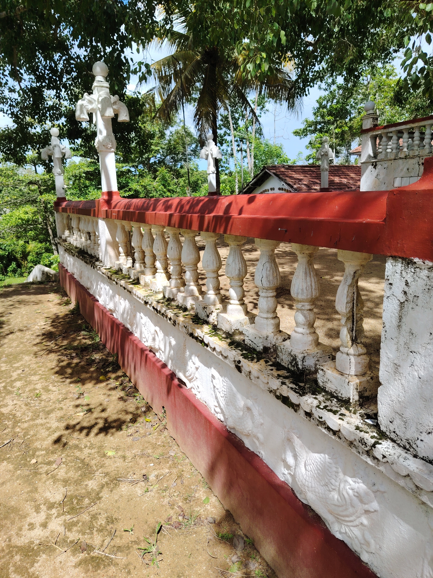 The image displays a decorative white and red balustrade or railing in an outdoor, sunlit setting. The railing is composed of numerous ornate white balusters, some of which exhibit cracks and dark moss/algae growth, indicating age and weathering. A vibrant red cap rail sits atop the balusters, complementing a matching red base along the bottom of the structure.
Below the balusters, a white wall section features intricate relief carvings of stylized bird-like figures, possibly peacocks or swans, arranged horizontally. This lower wall also shows signs of weathering. Interspersed along the balustrade are taller white posts, topped with spherical elements and adorned with stylized forms resembling animal or mythical creature heads.
In the background, dense green foliage and trees dominate the scene, including a prominent palm tree. Patches of clear blue sky are visible through the leaves, suggesting bright daylight. Further in the background, a building with a reddish-brown tiled roof can be glimpsed.
The foreground consists of a dirt or sandy ground with scattered small plants, leaves, and twigs. Strong, well-defined shadows from the overhead trees fall across the ground and parts of the railing, confirming it is daytime and sunny. No people are visible, and there is no discernible text. The scene conveys a tranquil, slightly aged atmosphere within a lush, natural environment, likely in a tropical or subtropical region.