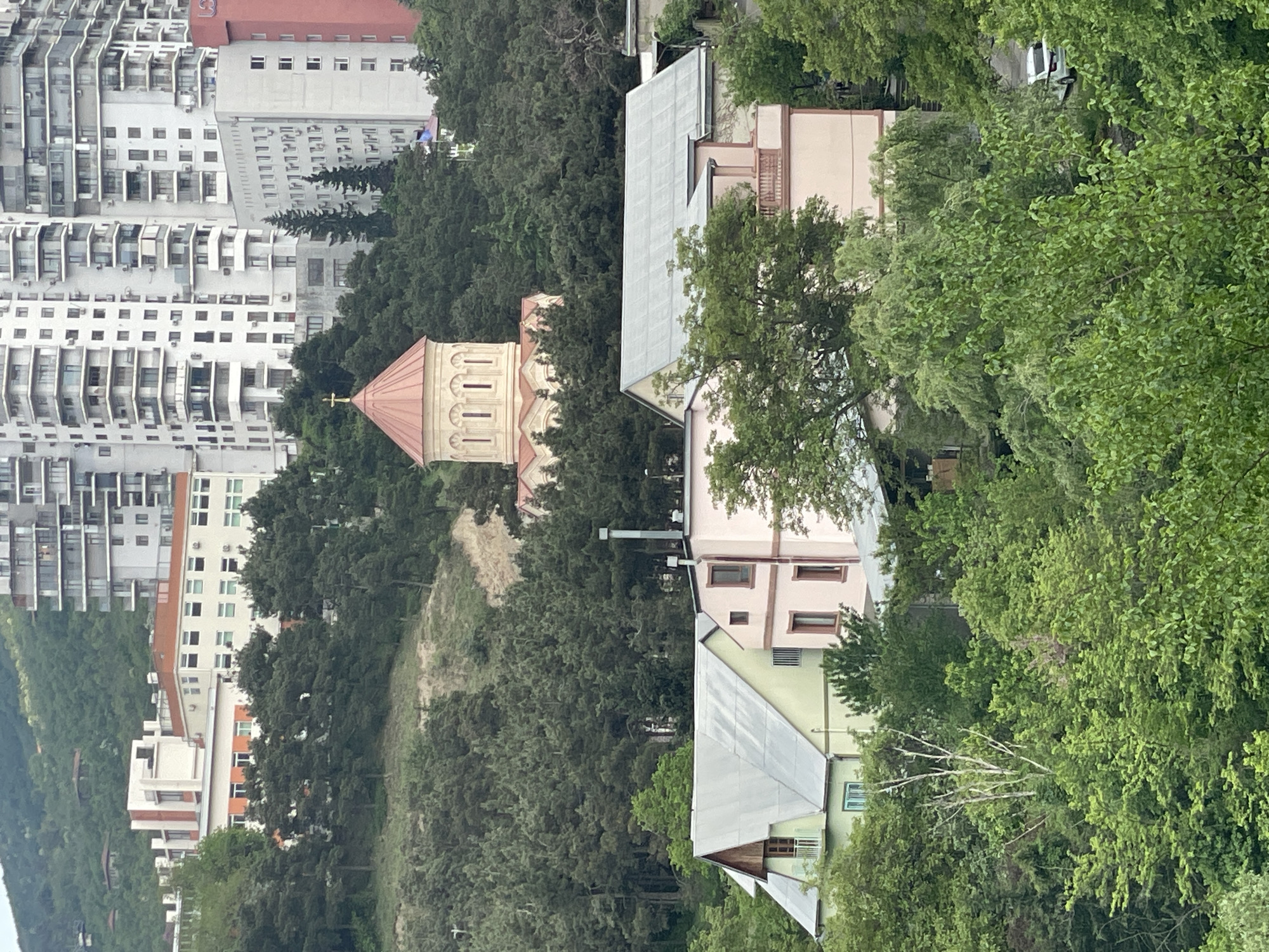 The image shows a view of Tbilisi, Georgia, with a mix of residential buildings, including high-rise apartments, and traditional houses with pitched roofs. A church with a prominent dome and cross sits among dense trees. The landscape features green hills and a variety of trees, suggesting a residential area with natural surroundings. The sky is not visible, indicating a daytime shot, possibly taken from an elevated position.