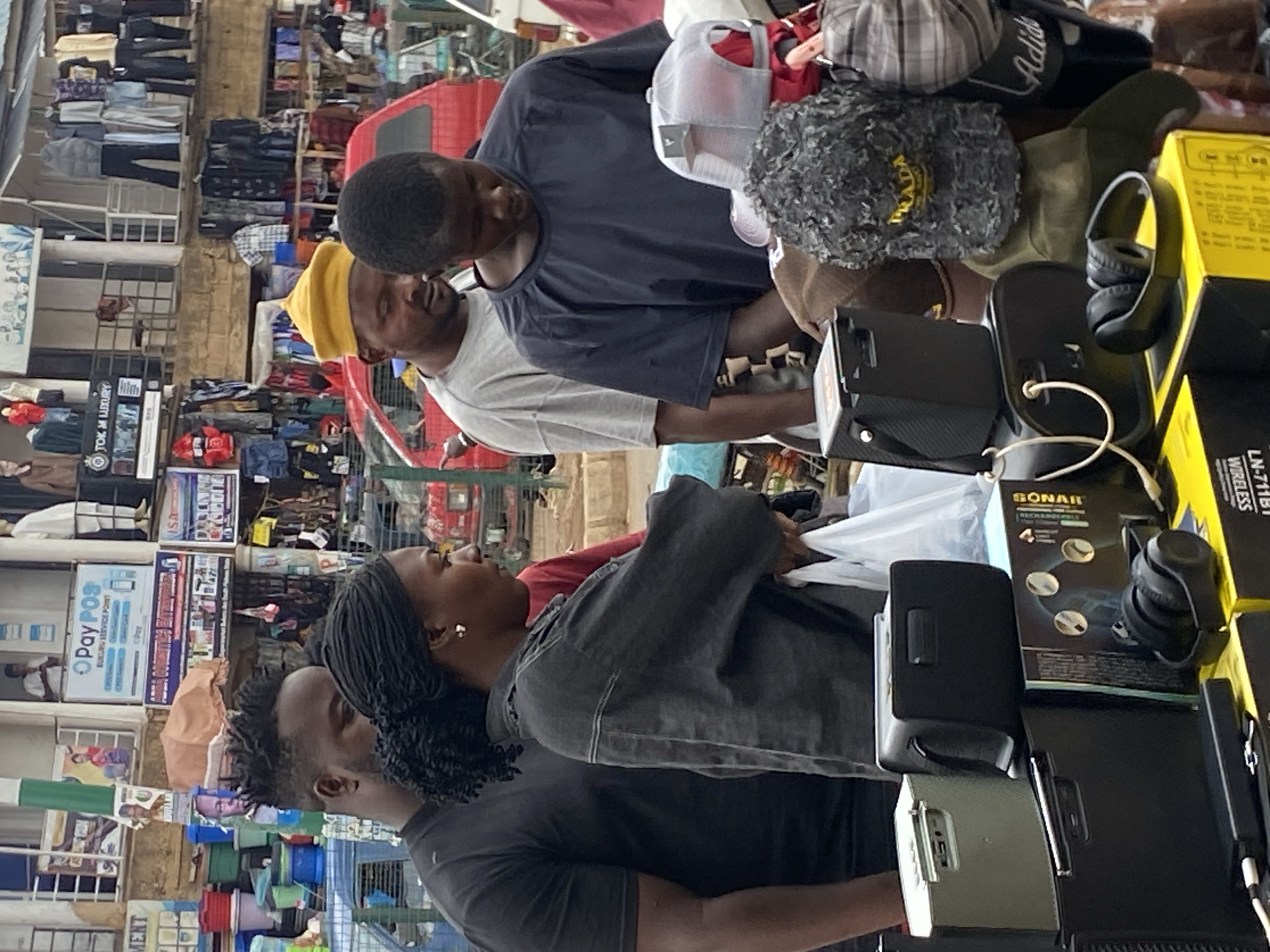 The image captures a vibrant and busy outdoor market scene in Bukuru, Nigeria, as indicated by a prominent sign. The overall setting is cluttered with merchandise and activity, typical of a bustling market environment.
In the foreground, several individuals are engaged with market stalls. To the right, two men are visible at a stall. One man, wearing a dark blue t-shirt, looks down at the items, while another man in a light grey t-shirt and a bright yellow beanie stands behind him, also observing the goods. Their stall displays a variety of items including hats (a grey textured hat with "PRADA" embroidered, a plaid cap, and a white mesh baseball cap), an "Adidas" slide sandal, black over-ear headphones, and several boxed electronic gadgets like speakers and power banks. Text visible on these boxes includes "GONAR", "SONAR", "WIRELESS", and "LN-711BI".
On the left side, two other individuals interact with another stall. A woman with dark braided hair, wearing a dark denim-like jacket, looks towards the left side of the frame. Behind her, a man with dreadlocks, dressed in a dark t-shirt, is partially visible.
The background features numerous other market stalls, packed with clothing on racks, shelves, and various other goods. Buildings line the market area, displaying signs. A prominent blue and white sign reads "OPay POS BUKURU SERVICE POINT," confirming the market's location. Below it, another sign reads "TOK M LUXURY." A red vehicle is partly visible in the mid-ground. The scene is illuminated by daylight, suggesting an active trading period.
