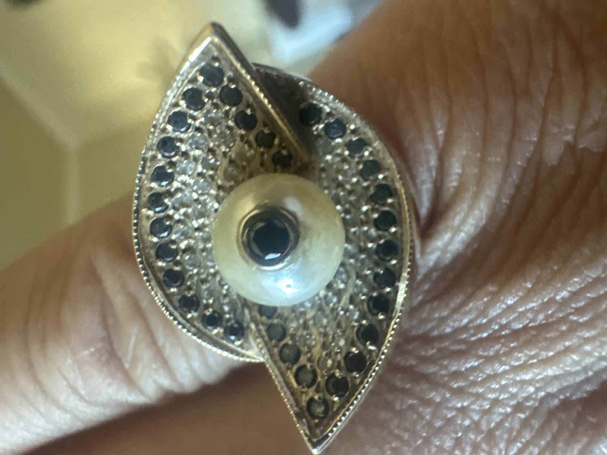 An eye-shaped ring with a pearl and black gemstones is worn on a finger. The ring features a central pearl surrounded by black stones and smaller clear stones. The finger is textured with skin creases. The background is out of focus and appears to be a pale yellow fabric. The image is a close-up shot. There is no visible indication of the location in Yakhroma, Russia.