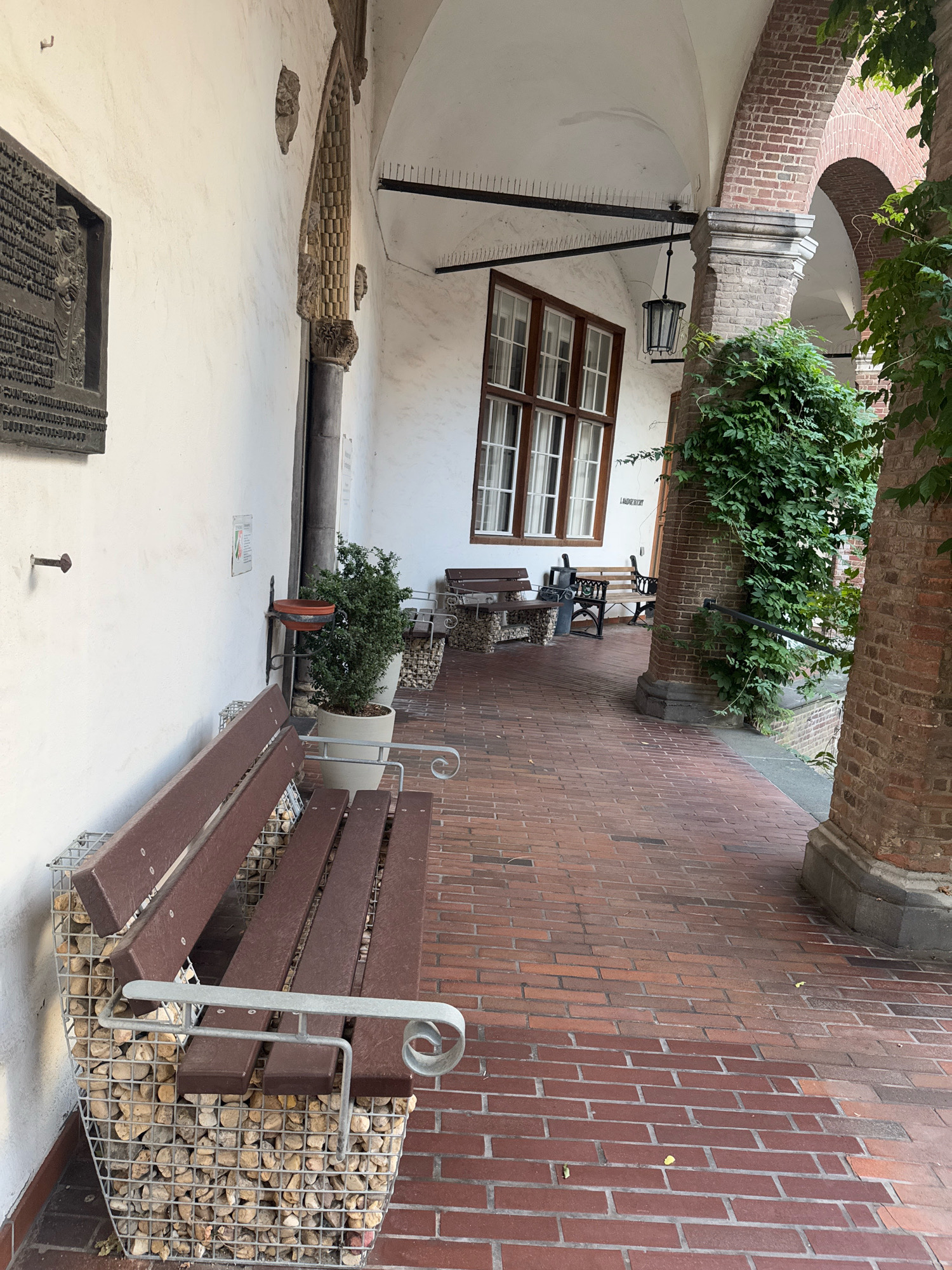 The image depicts a view down a covered walkway with red brick paving. On the left, a white wall features a dark plaque with relief text and an image, and a wall-mounted plant holder. A dark brown wooden bench with metal armrests sits in the foreground, its base constructed from a metal cage filled with stones. Further down the walkway, another bench is visible, along with a potted plant. The walkway is supported by brick arches, and the wall on the right is also constructed of brick. Green ivy grows along the brickwork. A large window with multiple panes is set into the white wall. The scene appears to be part of an outdoor public space in Cleves, Germany, likely within the grounds of a historical building or courtyard.