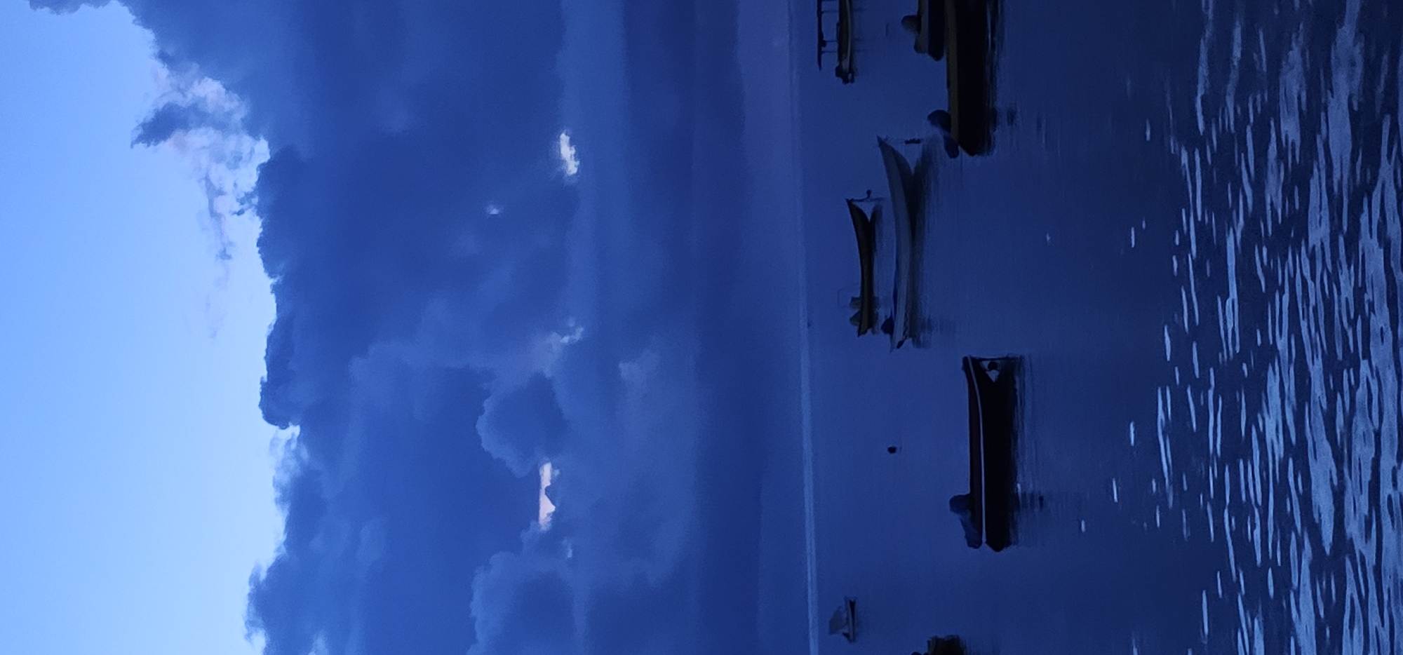 The image depicts a waterscape likely taken in or near Denpasar, Indonesia, judging from the style of the boats present. The sky on the left is partially obscured by dark, heavy clouds, some with breaks that reveal lighter sky. To the right of the clouds is the body of water, where several small boats are moored. The water is dark with visible ripples, reflecting the light of the sky. The boats appear to be traditional fishing vessels common in the region. The overall tone of the image is dark and suggests either an early morning or late evening scene.