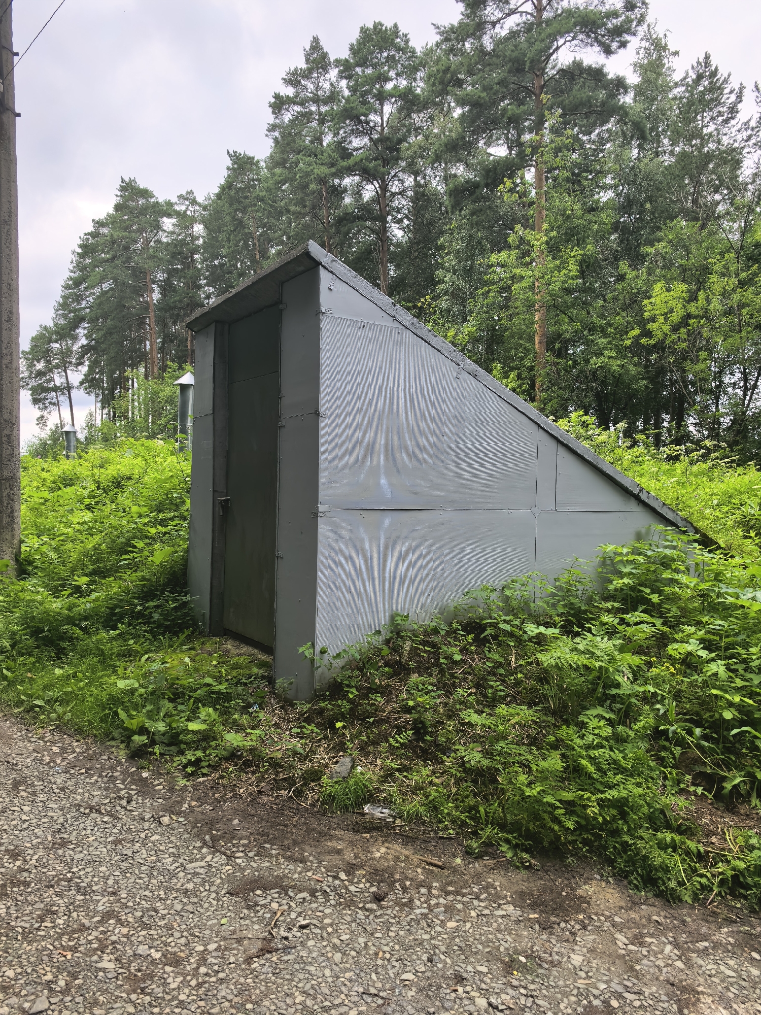 An exterior, eye-level, medium shot captures a grey, metallic, shed-like structure with a sloped roof, set amidst dense green foliage and tall pine trees. The shed has a single, dark grey door with a visible latch and hinges. The exterior of the shed is constructed from large, corrugated metal panels. To the left of the shed, a grey utility pole is visible. The ground in the foreground is covered with grey gravel. In the background, a dense forest of pine and deciduous trees creates a natural backdrop. The sky is overcast, indicating a cloudy day. The scene appears to be located in a natural or semi-natural area, possibly on the outskirts of Yekaterinburg, Russia, given the presence of pine trees commonly found in the region. There are no people or animals visible in the image.