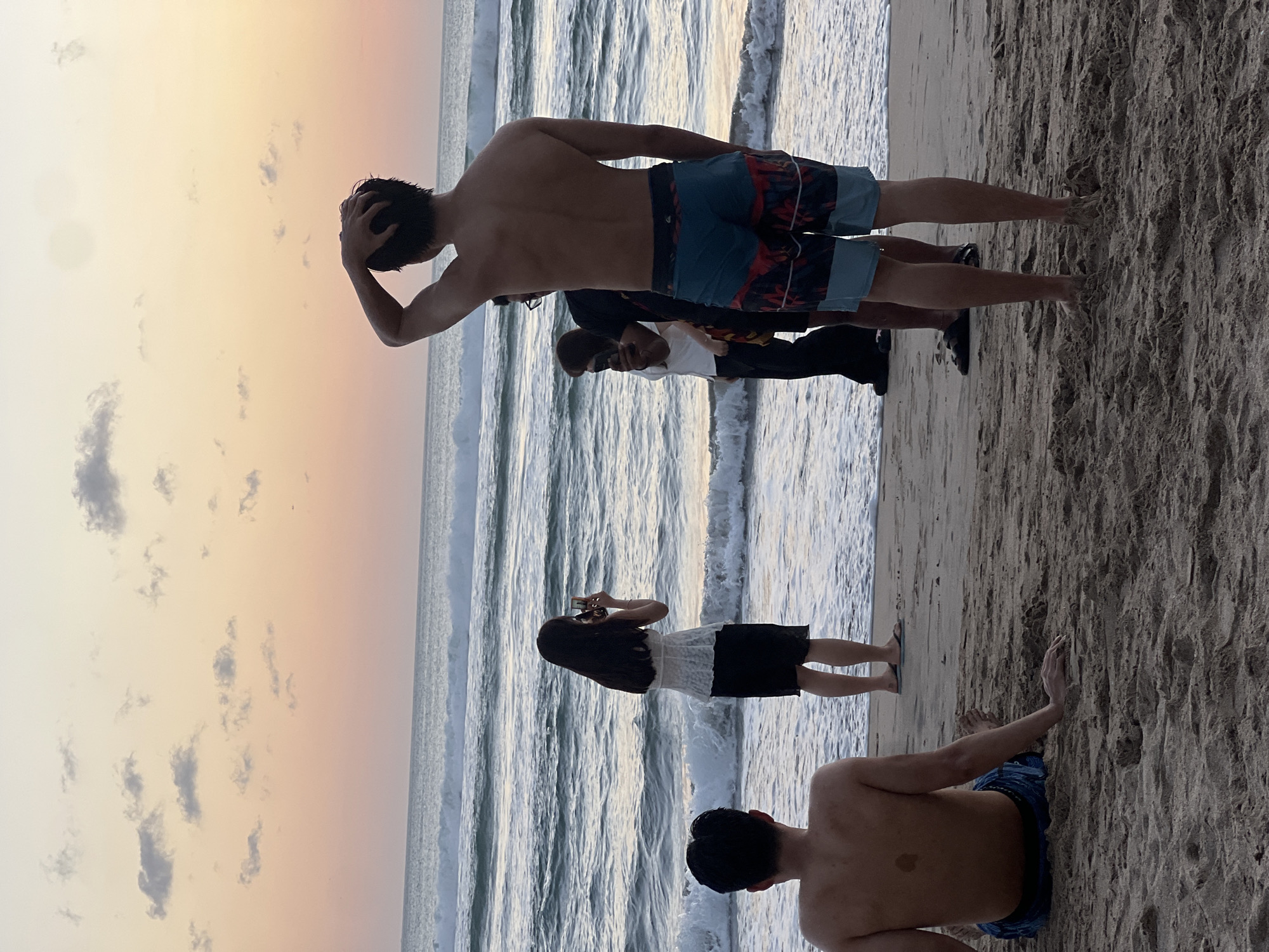 This image depicts a beach scene at sunset in Kuta, Indonesia. The sky displays a gradient of pastel colors, from soft orange near the horizon to pale yellow and pink higher up, with scattered gray clouds. The ocean is calm with gentle waves rolling towards the shore, and the water appears to be a muted blue-green.
In the foreground, on the sandy beach, two men are visible. One man is seated, facing away from the camera, with his back to the ocean and his left arm resting on the sand. He is wearing blue patterned swim trunks. The second man, also facing away, is standing closer to the water, with his right hand touching the back of his head. He is wearing colorful patterned swim trunks.
Further down the beach, near the water's edge, three people are gathered. A woman with long dark hair is standing and holding a smartphone, appearing to take a photo. She is wearing a white lacy top and a black skirt. Behind her, two individuals are standing and looking towards the ocean or at the woman. One person is wearing glasses and a dark t-shirt, and the other is wearing a white top. The interaction suggests casual observation or photography of the sunset or the surroundings. The overall atmosphere is relaxed and serene, typical of a beach at dusk.