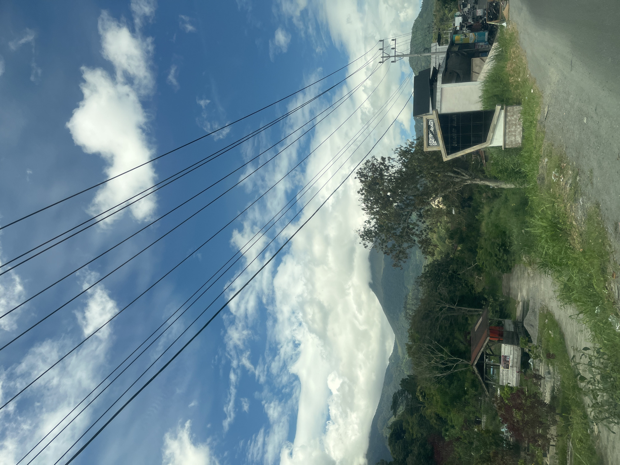 The image depicts a street scene in Simpanglayang, Indonesia, characterized by a road running through a verdant landscape. Overhead, a network of power lines crisscrosses the bright blue sky, dotted with fluffy white clouds. To the left of the road, a small, rustic wooden structure with a red roof sits amidst dense greenery. Further in the background, rolling hills and mountains, partially obscured by clouds, form a natural backdrop. On the right side of the road, a modern, angular building with a white facade and dark windows stands prominently. Adjacent to this building, a utility pole supports electrical wires. A few vehicles, including motorcycles, are visible parked or moving along the road. The overall impression is one of a rural or semi-rural setting within the city of Simpanglayang.