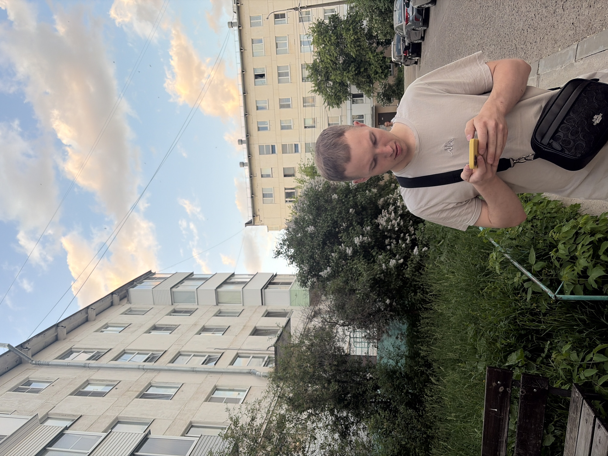 The image shows a young man in a beige t-shirt and black shoulder bag, holding a small yellow object. He is standing outside in an urban environment. Behind him, there are apartment buildings, green trees, and a patch of grass with a wooden bench. The sky is partially cloudy. The image appears to be taken in the city of Chaikovsky, Russia.