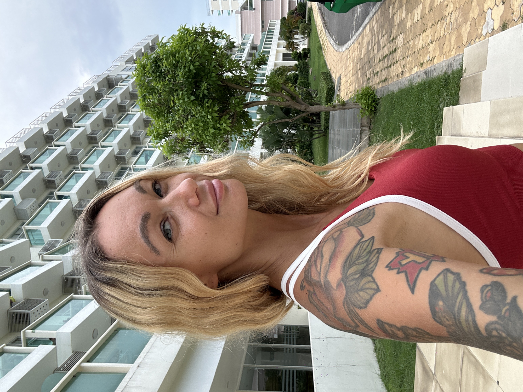 A blonde woman with visible tattoos on her left arm is posing for a selfie in front of a multi-story building. The woman is wearing a red sports bra. The building is white with many balconies and what appear to be air conditioning units. To the right of the building, there is a tree and a paved pathway. The background is overcast. The scene appears to be outdoors in a city, possibly Phu Thuy Ward, Vietnam, given the architectural style.