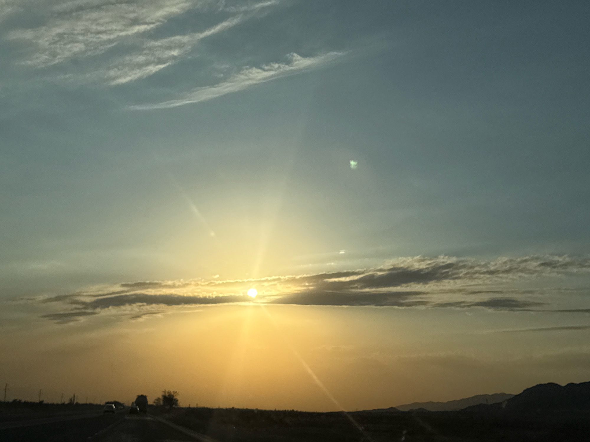 The image captures a serene sunset scene, with the sun positioned just above the horizon, casting a warm, golden glow across the sky. Wispy clouds, illuminated by the fading light, stretch across the expansive sky. The foreground is dominated by a dark, flat terrain, possibly a plain or desert, with faint outlines of distant hills or mountains on the horizon. A road, likely a highway, is visible in the lower part of the image, with a few cars driving along it. The overall atmosphere is tranquil and peaceful, evoking the beauty of a natural landscape at dusk. The location is identified as being in the city of Chüy, Kyrgyzstan.