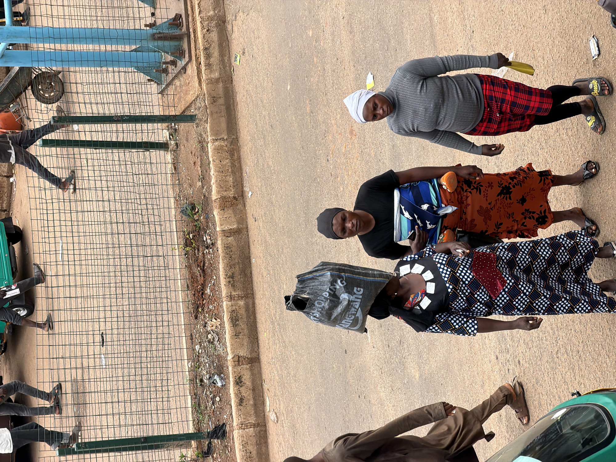 This is an outdoor, eye-level, medium shot photograph taken in Bukuru, Nigeria. In the foreground, three women are standing on a paved road. The woman on the left is wearing a black headscarf, a black t-shirt, and an orange and black floral skirt. She is carrying a child wrapped in a blue and white patterned cloth on her back. The woman in the middle is wearing a black headscarf and a black t-shirt, and an orange skirt with a floral pattern. The woman on the right is wearing a white headscarf, a grey turtleneck sweater, and a red and black plaid skirt. She is holding a yellow object in her right hand.
Behind the women, a black plastic bag with the words "SHOPPING / ANG WANG" printed on it is balanced on the head of another person who is partially visible from the shoulders up and facing away from the camera. This person is wearing a black and white polka-dot dress with a red rectangular accent and a black patterned scarf.
Further in the background, a metal fence separates the road from a dirt area with some debris. Beyond the fence, several figures are walking, and the wheel of a vehicle, possibly a tricycle or a truck, is visible. A blue metal structure, likely a support pole or pillar, is also present in the background. The overall environment suggests a public street or marketplace in an urban setting.