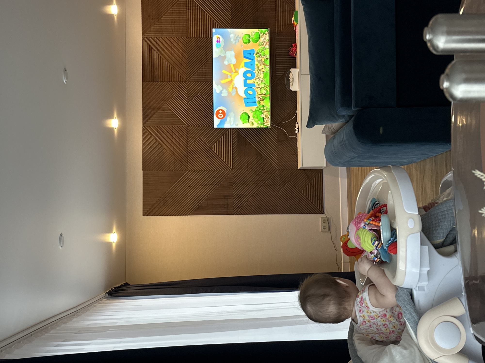The image depicts an interior scene. The walls are painted white with recessed lighting fixtures. One wall is decorated with a geometric, textured, brown panel. A TV is mounted on this panel, displaying a cartoon. A dark blue sofa is partially visible. A white baby highchair with toys is also present, occupied by a baby, who is facing away from the camera and interacting with the toys. The window has white curtains and a black valance. The picture was taken indoors, perhaps in an apartment in Подшивалово, Russia.