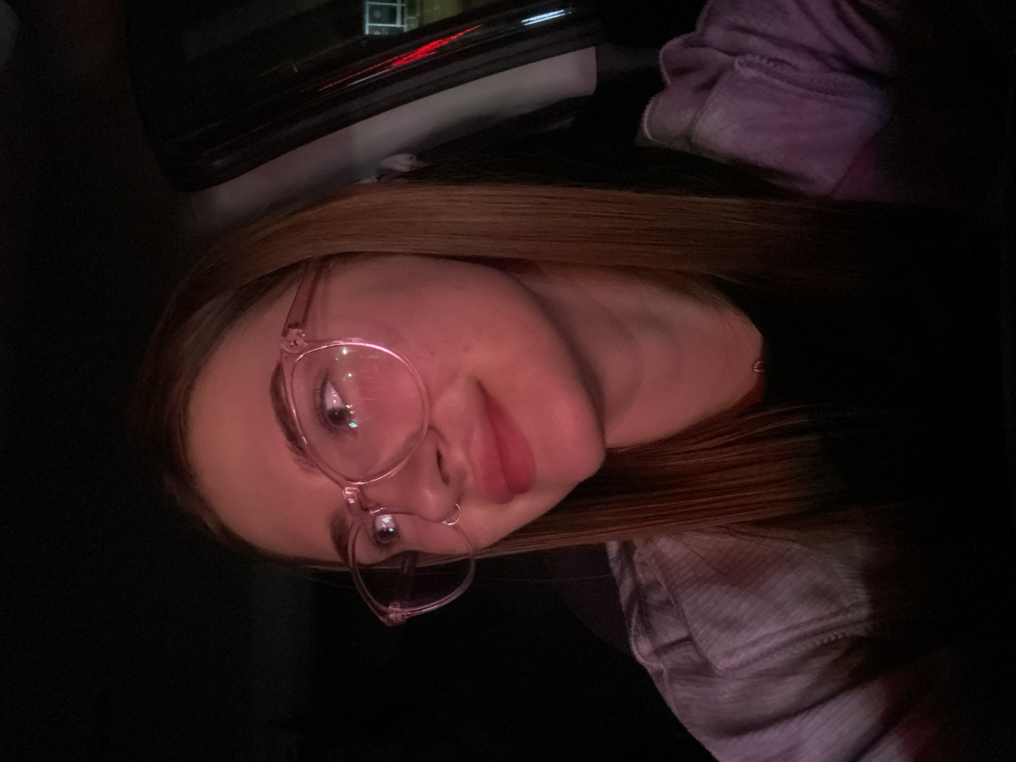 The image shows a young woman with long, straight, reddish-brown hair and light skin, wearing large, clear-rimmed glasses. She is looking to her left with a slight smile. She is wearing a dark shirt under a light-colored, textured garment, possibly a jacket or sweater, which is dimly lit and reflects pink and purple hues. A subtle necklace is visible around her neck. The background is dark, with hints of what appears to be a car interior, including a portion of a car door and possibly a window with distant lights visible through it. The lighting suggests the photo was taken at night or in a dimly lit environment. There is no information in the image that would allow for a specific location within the city of Dyakovo, Russia.