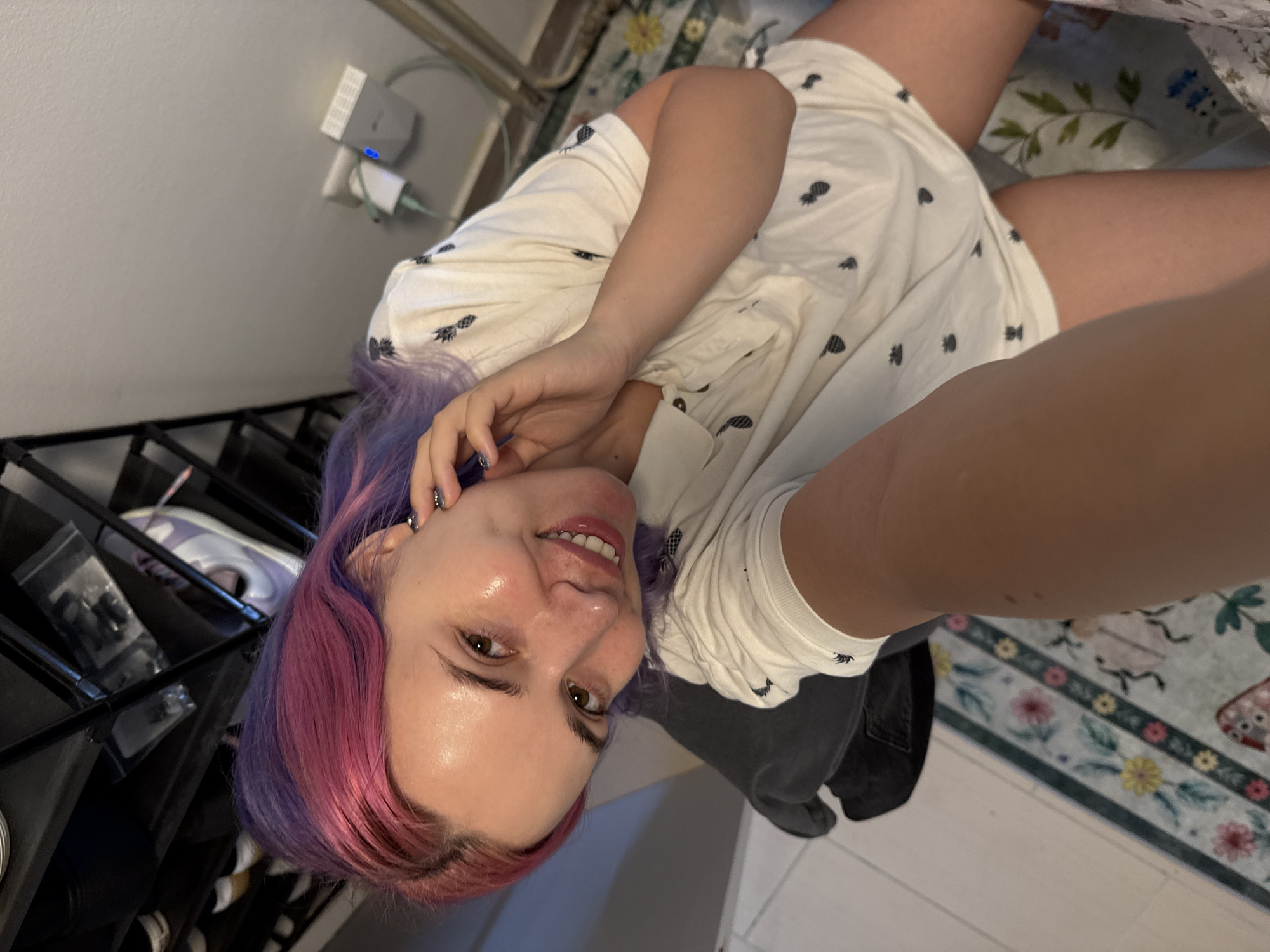A selfie depicts a woman with dyed purple and pink hair, wearing a white pineapple-patterned t-shirt. She is indoors, and a black metal shelving unit is visible to the left. A rug with floral designs is present on the floor to her right. The presence of an electrical outlet and a network device suggests a residential setting. The city of City of Zagreb, Croatia, cannot be determined from the image.