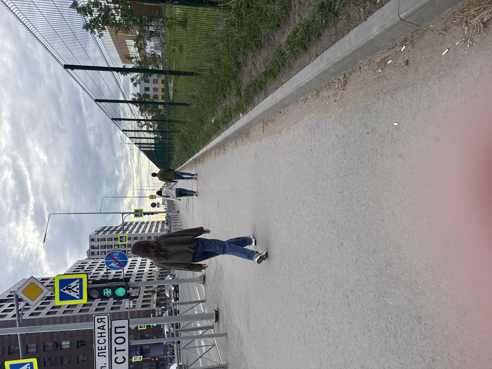 The image depicts a sidewalk scene in the city of Perm, Russia, likely in a newly developed urban area. In the foreground, a woman with long, dark hair wearing a dark green sweater and blue jeans walks away from the camera. She is wearing white sneakers. To her right, a young person in a white hoodie and blue jeans also walks away, followed by another individual in a green jacket and dark pants. They are all on a paved sidewalk adjacent to a road with a metal railing and parked cars visible in the background. Traffic lights and various road signs, including a pedestrian crossing sign and a "STOP" sign, are present along the roadside, indicating an intersection. A tall green metal fence runs along the right side of the sidewalk, separating it from a grassy area and modern buildings. The sky is overcast with a mixture of gray clouds and a hint of diffused sunlight. The overall scene suggests an urban environment in Perm, possibly during the evening or a cloudy day.