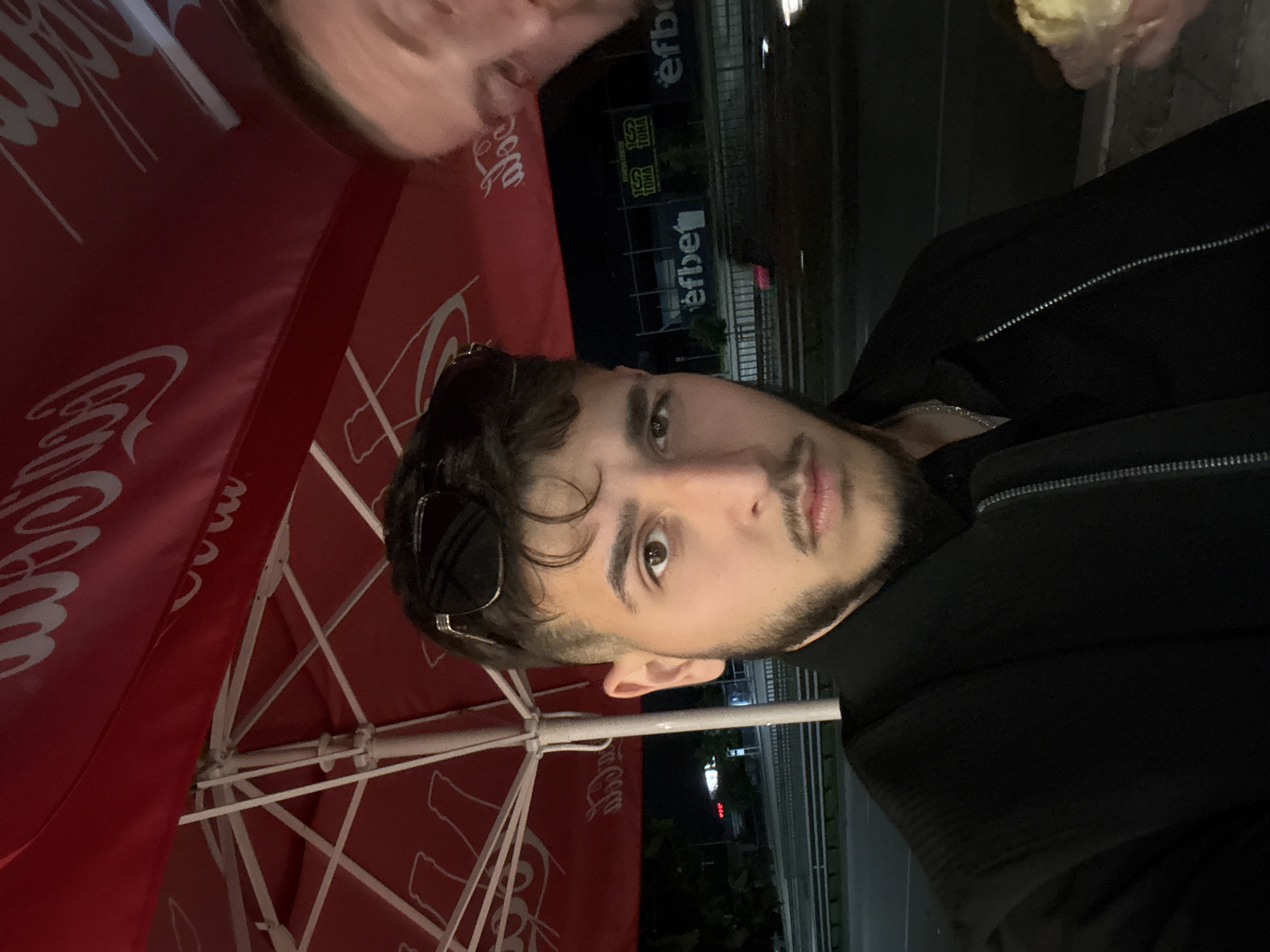 A young adult male with dark hair, a trimmed beard, and a mustache is prominently featured in the left and center of the image, looking directly at the camera with a neutral expression. He wears sunglasses pushed onto his head and a black collared jacket with a zipper, revealing a silver chain around his neck. To his right, the blurred forehead and partially visible face of another person are seen, holding a light-colored food item in their hand at the bottom right. The scene is outdoors at night, under two red umbrellas displaying white "Coca-Cola" branding. In the background, across a multi-lane boulevard, an illuminated white "efbet" sign is visible on a building, while further left and slightly behind, a green and yellow sign for "100 ТОНА GYM" is displayed on another building. A metal barrier separates the seating area from the boulevard.
The location is identified as Slivnitsa Boulevard in Varna, Bulgaria, specifically near the intersection with the street leading to the Sports Hall. The "efbet" sign visible across the street corresponds to the efbet casino at Slivnitsa 172, and the "100 ТОНА GYM" sign identifies the "100 ТОНА Fitness & Spa" at Slivnitsa 170. The individuals are seated at an outdoor establishment, likely a cafe or kiosk, on the sidewalk adjacent to Slivnitsa Boulevard.