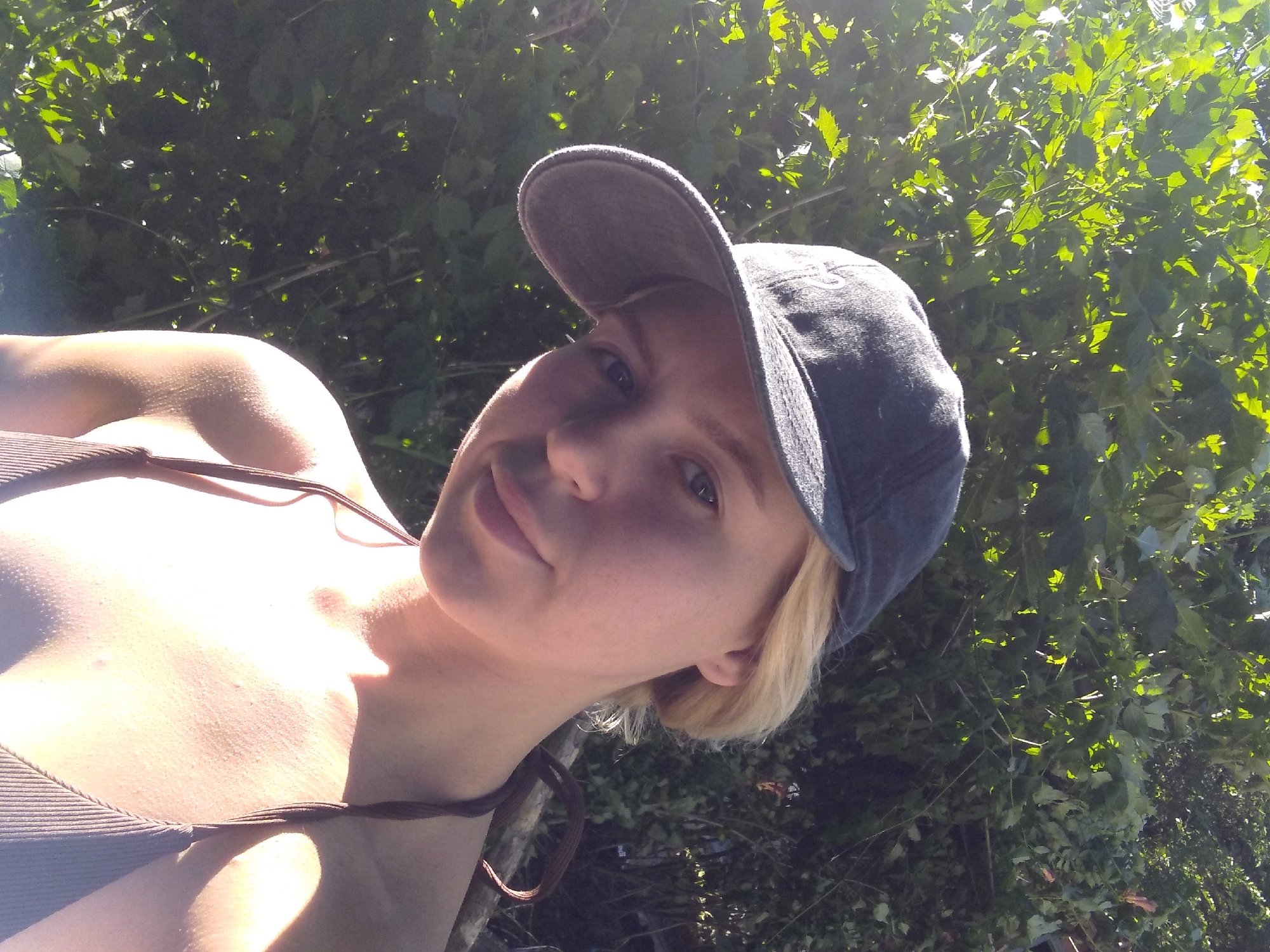 A young woman wearing a dark baseball cap and a ribbed brown tank top is shown in a close-up, angled selfie. Her fair skin is illuminated by bright sunlight, casting strong shadows and highlights. She has blonde hair and light-colored eyes, with a slight smile. The background is filled with lush green foliage, suggesting an outdoor setting. The leaves are dappled with sunlight, indicating a sunny day. The overall impression is casual and relaxed, likely taken outdoors on a bright day. There is no visible text in the image. The image appears to be taken in Radisne, Ukraine.