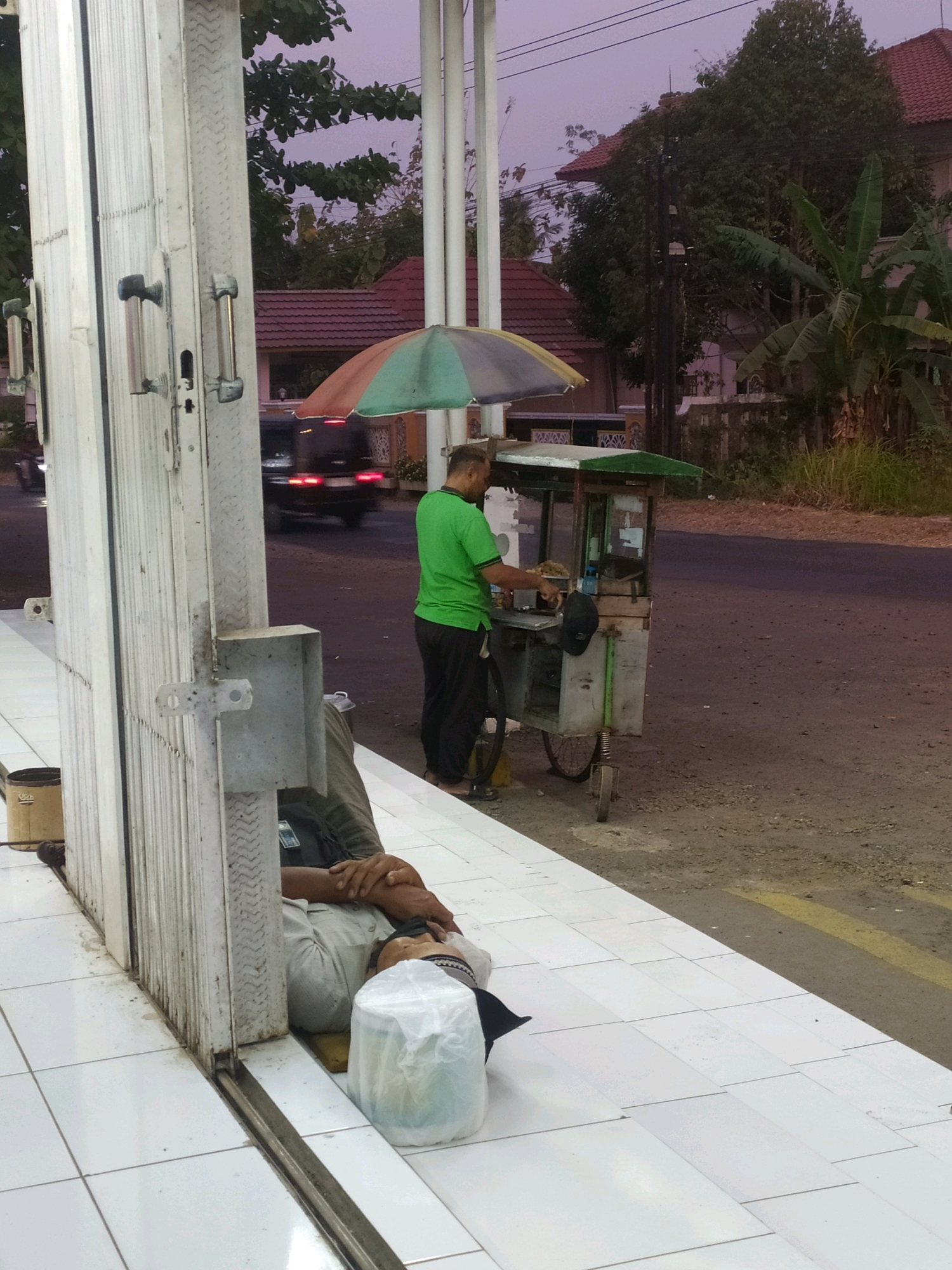 The image depicts an outdoor scene at dusk, likely in Tambaksogra, Indonesia, characterized by an overcast sky. In the foreground, a person is lying down on a white tiled surface, resting against a white corrugated metal gate. They are covered by a light-colored garment and have a white plastic bag next to their head. To the right of the gate, a street vendor is operating a cart with a multi-colored umbrella. The vendor, wearing a green shirt and dark pants, appears to be serving a customer or attending to their cart. The cart has wheels and appears to be mobile. Behind the vendor, a black vehicle is visible on the asphalt road, its taillights illuminated. The background shows vegetation, including trees and what appear to be banana plants, and houses with red tiled roofs, suggesting a residential area. The overall atmosphere is tranquil, with the fading light indicating the end of the day.