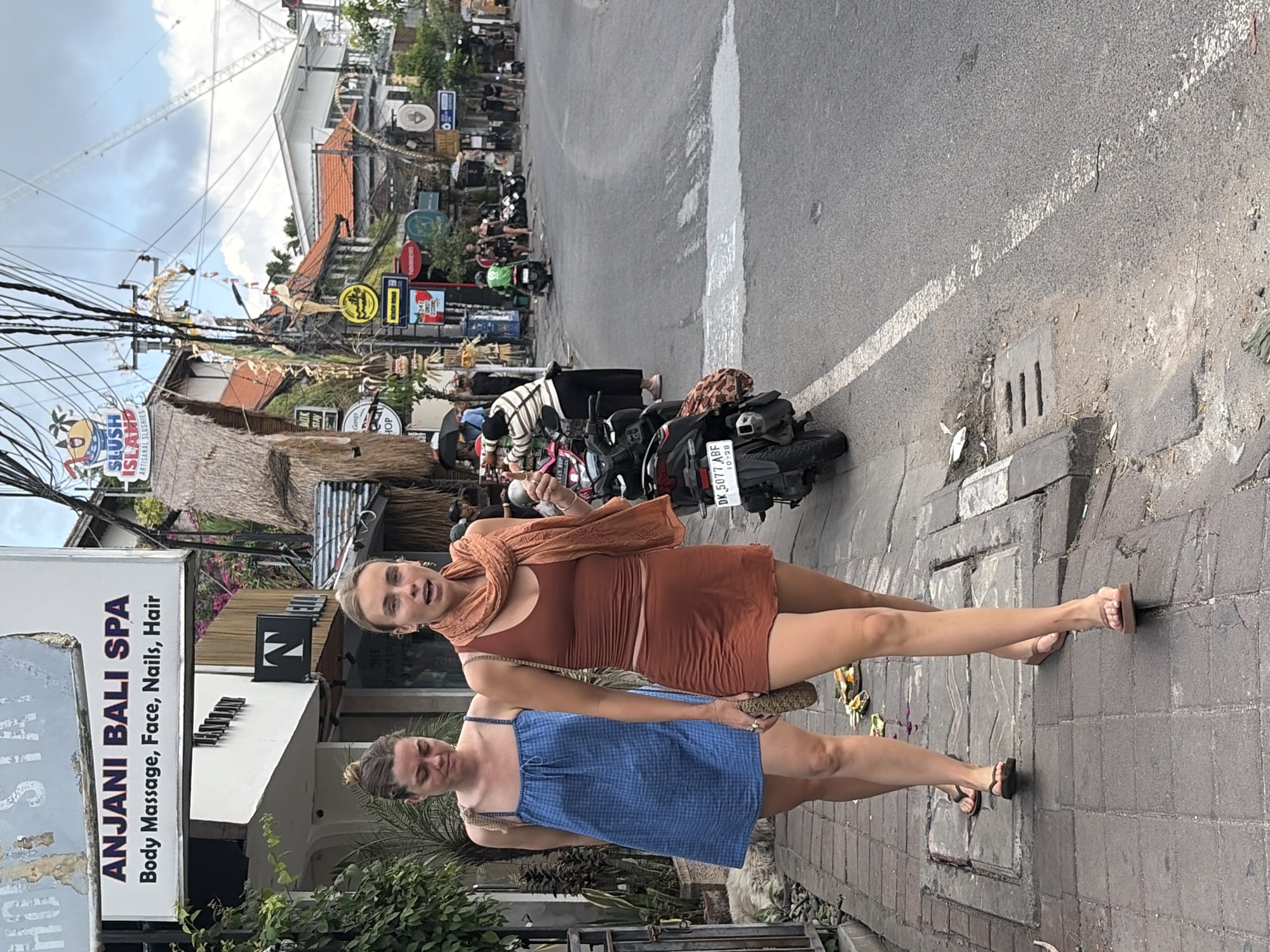 Two young women walk along a paved sidewalk next to a street in North Kuta, Indonesia. The woman on the left wears a blue checkered dress and flip-flops. The woman on the right wears a brown tank top, a brown mini skirt, a rust-colored scarf, and flip-flops. She is speaking and gesturing with her left hand. Behind them, a black motorcycle is parked on the side of the road. The street is lined with shops and palm trees, and a power line runs overhead. A sign for "ANJANI BALI SPA" is visible on the left side of the image. The street is busy with other pedestrians and motorcycles in the background.