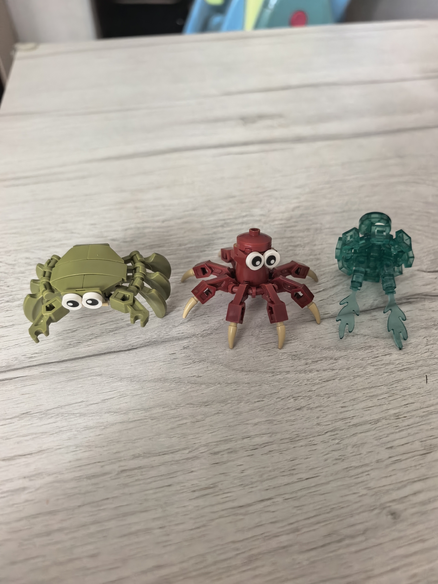 Three toy creatures, seemingly constructed from building blocks, are arranged in a row on a light-colored wooden surface. The surface displays a distinct wood grain pattern.
On the left is an olive green, crab-like figure. It features a segmented shell and multiple jointed legs, along with prominent white googly eyes that give it a whimsical appearance.
In the center is a dark red, spider-like creature. It has a round body and eight jointed legs that end in tan, pointed tips. This figure also sports a pair of white googly eyes.
On the right is a translucent teal-colored figure with a more abstract, perhaps aquatic, design. It has a somewhat angular body and two large, fin-like or tentacle-like appendages extending backward.
The creatures are static, appearing to be posed for display or a photograph. The background is softly blurred, showing out-of-focus elements that suggest an indoor setting, possibly another toy or object with bright colors visible in the upper part of the frame. There is no visible text in the image. The scene is well-lit, characteristic of daylight or bright artificial lighting indoors. The image was captured in Sterlitamak, Russia.