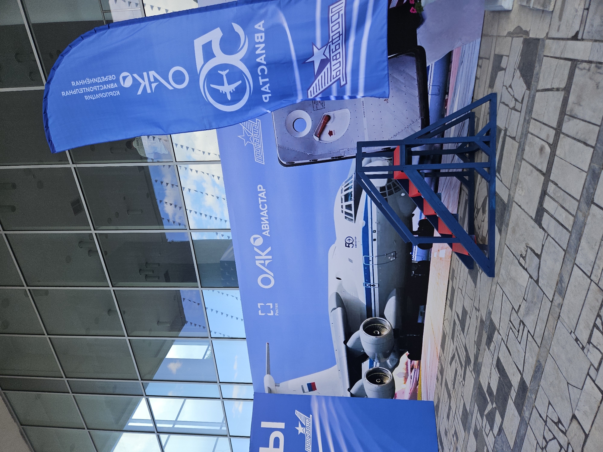 The image shows an outdoor exhibition space in Ulyanovsk, Russia, with a large banner featuring an airplane and company logos. The banner is blue and white, with the text "OAK" and "Aviastar" visible. A large airplane is depicted on the banner, along with the number "50" and a logo of an airplane inside a circle. To the right of the banner, a blue flag with white text and logos is standing upright. In front of the banner, a section of a real airplane door is displayed, and a blue metal structure with red steps is positioned in front of it, possibly for visitors to access the display. The ground is paved with stone tiles. The background features a modern building with large glass windows that reflect the sky and surroundings. No people are clearly visible in the frame, though a blurry figure can be seen in the reflection of the windows.