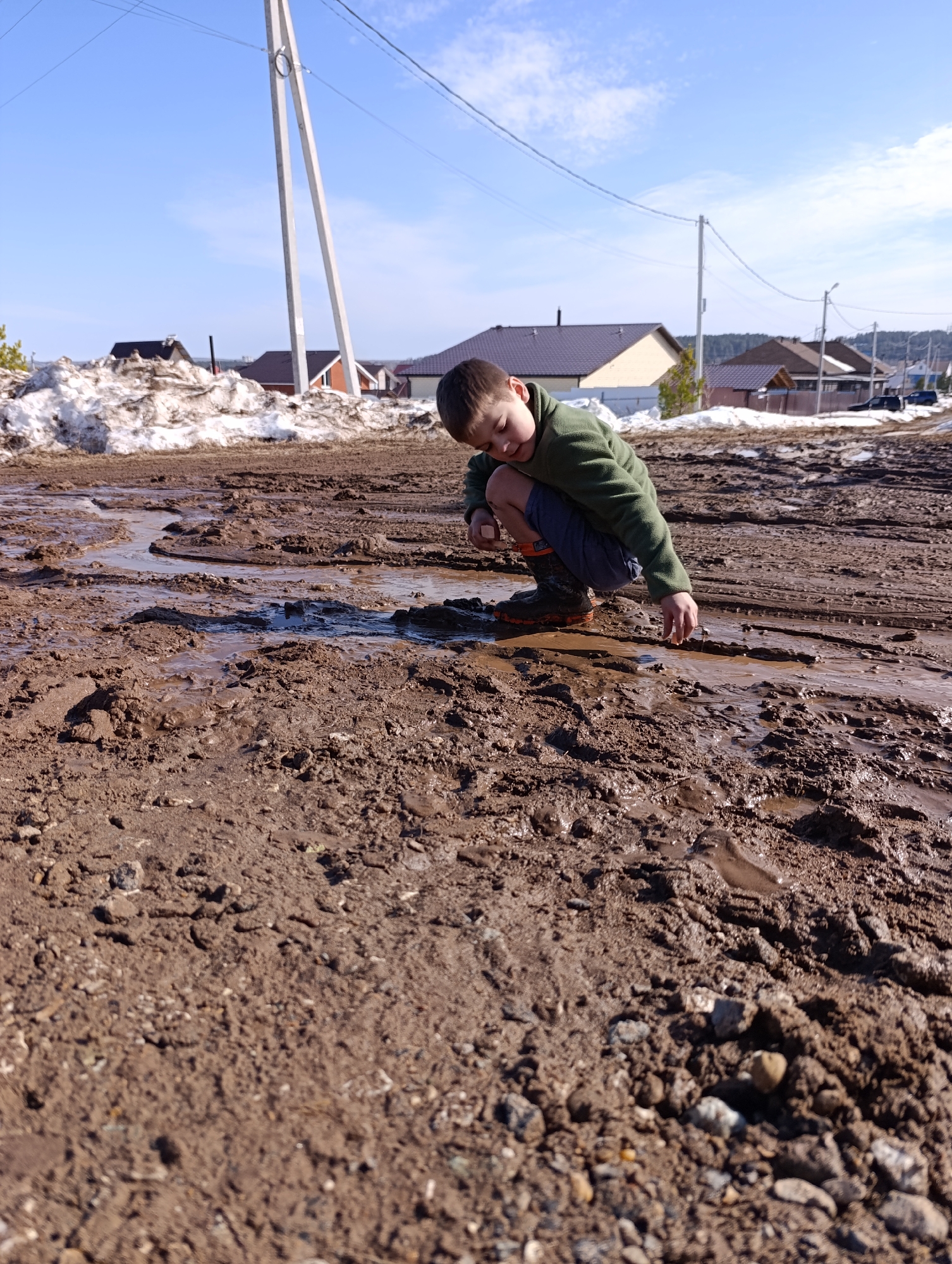 A young male, possibly a child, is depicted crouching on an unpaved, muddy surface. He wears a green long-sleeved top, dark shorts, and dark, possibly waterproof, boots with orange accents. His head is angled down towards the ground, and he appears to be interacting with a small pool of water or wet mud with his right hand. The ground is heavily disturbed, covered in dark brown mud with visible tire tracks, footprints, and numerous puddles of water reflecting the sky. In the mid-ground, to the left, is a large pile of melting snow. The background features several residential houses with varying roof colors (dark grey, orange-brown) and construction styles, characteristic of a suburban or semi-rural area. Multiple utility poles with overhead power lines are visible against a clear blue sky with sparse white clouds. This scene is located in a residential area of Chaikovsky, Russia, during what appears to be the spring thaw.