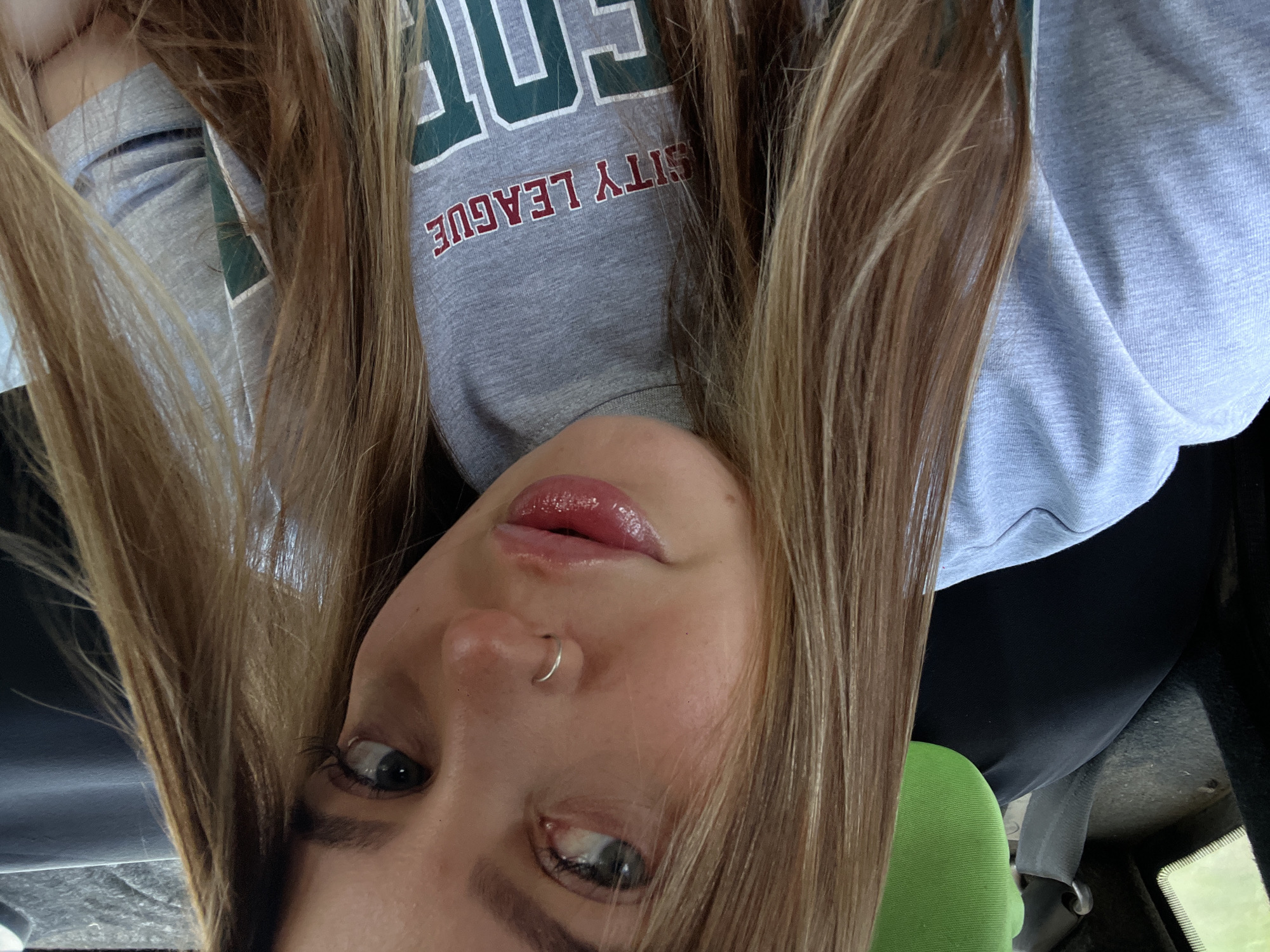 The image presents a close-up, slightly upward-angled portrait of a young adult female, likely captured as a selfie from within a vehicle. The subject has long, multi-toned hair, predominantly light brown with blonde highlights, flowing over her shoulders and framing her face. She has light-colored eyes, appearing blue or green, directed slightly upward and to her left. A small, silver hoop piercing is present in her left nostril, and her full lips are slightly parted, coated with a glossy, pinkish-red product.
She is dressed in a light grey crew-neck garment, possibly a t-shirt or sweatshirt. Across the chest, the text "SITY LEAGUE" is visible in red lettering, followed by larger green block letters outlined in white, which appear to spell "FOR" or "COR".
The background indicates an interior of a vehicle. To the upper left, a dark seatbelt strap and buckle are discernible against a dark interior panel. A small reflective surface or window shows a blurred green color beyond. Behind the subject's head, a prominent, bright green, textured fabric object, likely a headrest cover or pillow, is visible. The right side of the background is darker and less defined, consistent with a vehicle's interior.
The visual information contained within the image does not provide any discernible identifying markers or cues to confirm that the scene is situated in the city of Novopokrovskaya, Russia.
