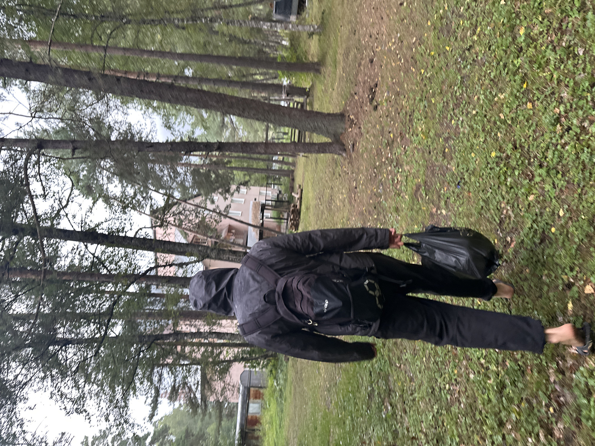 A person walks away from the camera in a wooded area. The person is wearing a black hooded jacket, black pants, and sandals, and is carrying a black backpack and a black plastic bag. The ground is covered in grass and fallen leaves, and the area is populated by tall, slender trees. In the background, there are buildings and a fence. The overall scene suggests a casual outdoor setting, possibly a park or residential area with natural surroundings. The image does not contain any elements that can identify the location as Arkhyz, Russia, or any specific city.