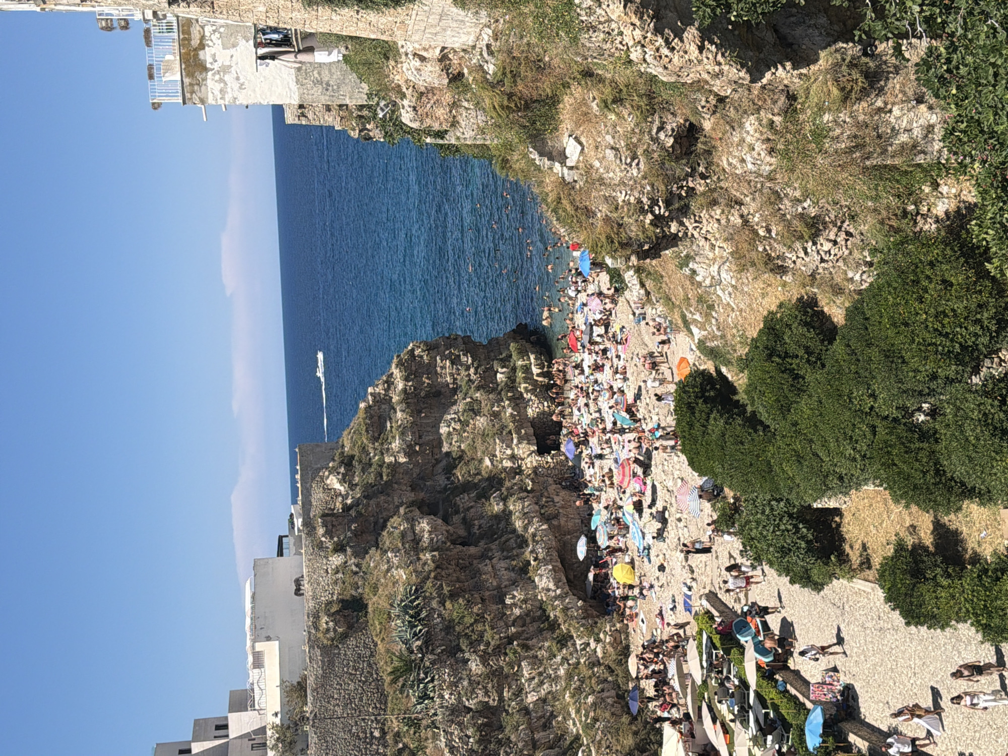 A high-angle, sun-drenched, midday view captures a crowded beach nestled within a cove in Polignano a Mare, Italy. The pebble beach is packed with people sunbathing under a multitude of colorful umbrellas, while others swim in the clear, blue Mediterranean Sea. Rocky cliffs, sparsely covered with vegetation, flank the cove, with buildings perched on the top of the cliffs. A white boat is visible in the distance on the water. The foreground features a paved walkway lined with lush green bushes and more people strolling along. The scene is bustling with activity, indicative of a popular tourist destination during peak season.