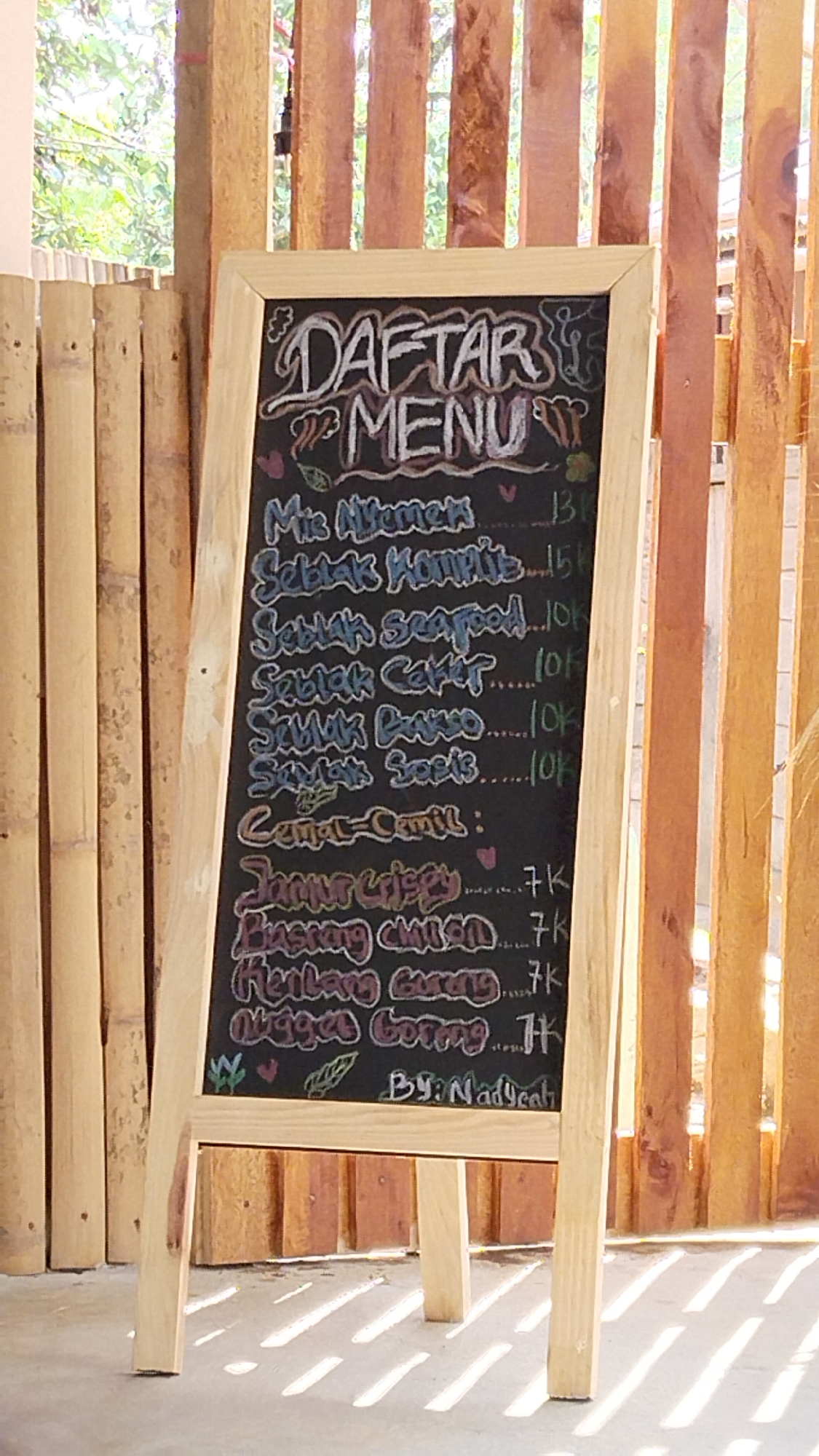 A wooden easel blackboard displays a menu written in chalk. The blackboard has a wooden frame and is set up outdoors, possibly in a cafe or street food stall. The background consists of vertical wooden planks, and some bamboo poles are visible on the left side. The sun casts striped shadows on the ground in front of the blackboard.
The menu is written in Indonesian. At the top, it reads "DAFTAR MENU" which translates to "MENU LIST". Below that are several food items with prices:
* Mie Nyemek - 13k
* Seblak Komplit - 15k
* Seblak Seafood - 10k
* Seblak Ceker - 10k
* Seblak Bakso - 10k
* Seblak Sosis - 10k
Then there is a section titled "Cemal-Cemil:" which means "Snacks:". Under this are:
* Jamur Crispy - 7k
* Singkong Goreng - 7k
* Nugget Goreng - 7k
At the bottom of the menu, it says "By: Nadyeah".
The image is taken from a medium-low angle, focusing on the blackboard. The lighting suggests it is daytime, with sunlight filtering through the background. No people are visible in the image. The overall scene depicts an informal food establishment. The mention of "Margatunggal, Indonesia" in the prompt suggests this location, but this information cannot be visually confirmed from the image itself.