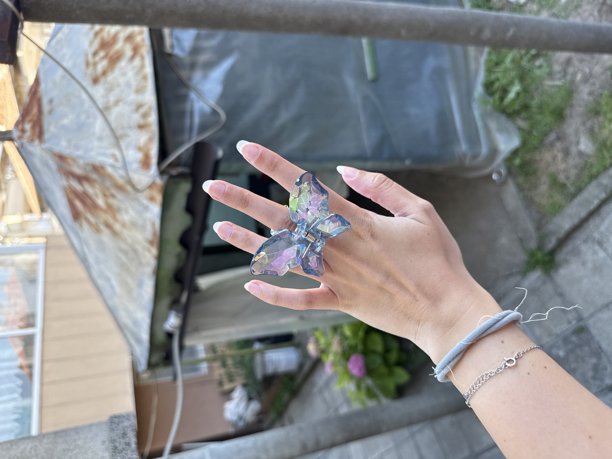 An outdoor, eye-level, medium shot captures a person's hand adorned with a clear, iridescent butterfly-shaped hair clip. The hand, with neatly manicured nails, is positioned in the center of the frame, showcasing the accessory. The background is blurred but suggests an urban setting, possibly a backyard or alleyway, with hints of vegetation and structures. In the distance, a rusty metal roof and a tarp-covered structure are visible, along with a building with a light-colored facade and windows. The scene's lighting is natural, indicating daylight. The image was taken in Tsarevo, Bulgaria.