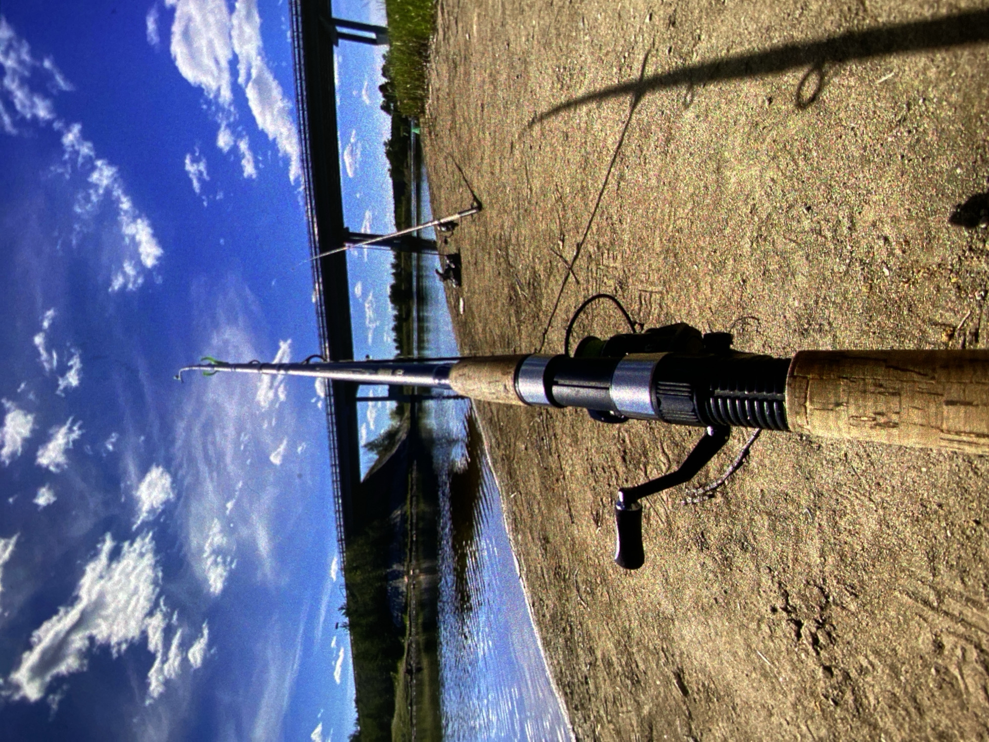 The image captures an outdoor scene, likely a fishing spot along a river or canal bank in Saint Petersburg, Russia, under a bright blue sky.
In the foreground, a fishing rod with a cork handle and a black reel is prominently positioned on the sandy, gravelly ground. The rod extends towards the right side of the frame. The textured ground, composed of small stones and dirt, shows a long, dark shadow of an unseen object or person, indicating bright sunlight.
Further back and to the left, another fishing rod is visible, set up with a stand on the bank, with its line extending into the calm water. The water body itself reflects the clear blue sky and scattered white clouds. The opposite bank features a line of green trees and foliage.
In the background, a large bridge structure spans across the water. Its dark pillars and framework are silhouetted against the vivid blue sky, which is adorned with numerous white cumulus clouds. The sky occupies a significant portion of the upper frame.
The overall scene conveys a sense of tranquility and leisure, characteristic of a day spent fishing. The time of day appears to be daytime, likely late morning or afternoon, given the strong shadows and clear sky with fair weather. No individuals are visible in the image, and there is no discernible text on any of the objects or in the environment.