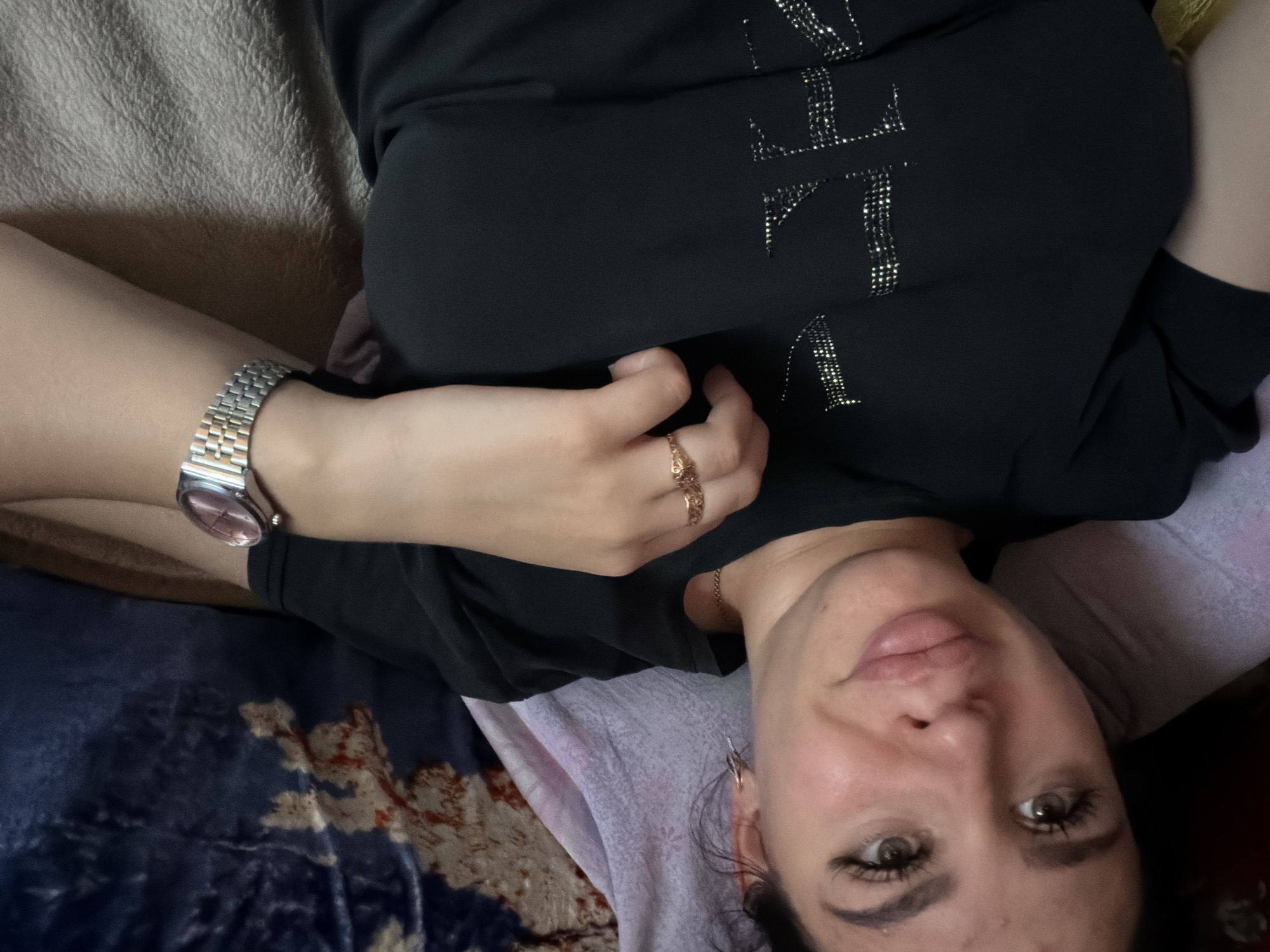 A young woman with dark hair, full lips, and prominent eyelashes is lying down, looking at the camera. She is wearing a black t-shirt with a silver, sparkly design on it, a gold ring, and a silver watch on her left wrist. The background includes a patterned blanket and a textured surface that appears to be a bed or couch. The image was taken indoors, likely in a residential setting. There is no indication of the specific location being in the city of Большой Букор, Russia.