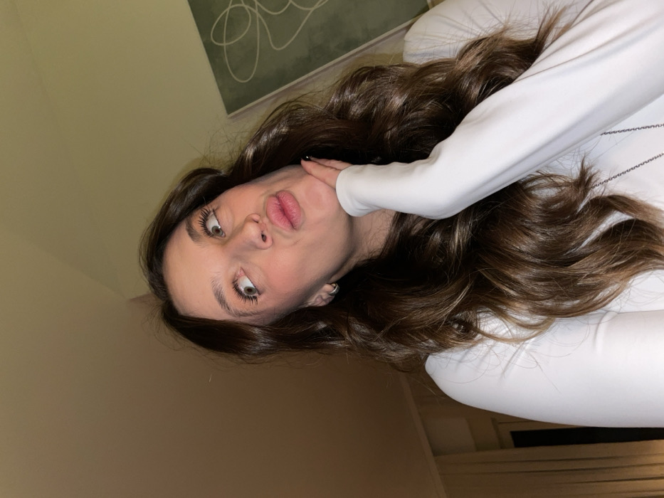 A young woman with long, wavy brown hair and light skin lies on her back, posing for the camera with a comical expression. Her lips are puckered, and her eyes look upwards. She wears a white, long-sleeved top. Her left hand is placed near her face. The background includes a wall, a piece of artwork, and a white pillow or surface. The image is slightly tilted, adding to the playful and candid feel.