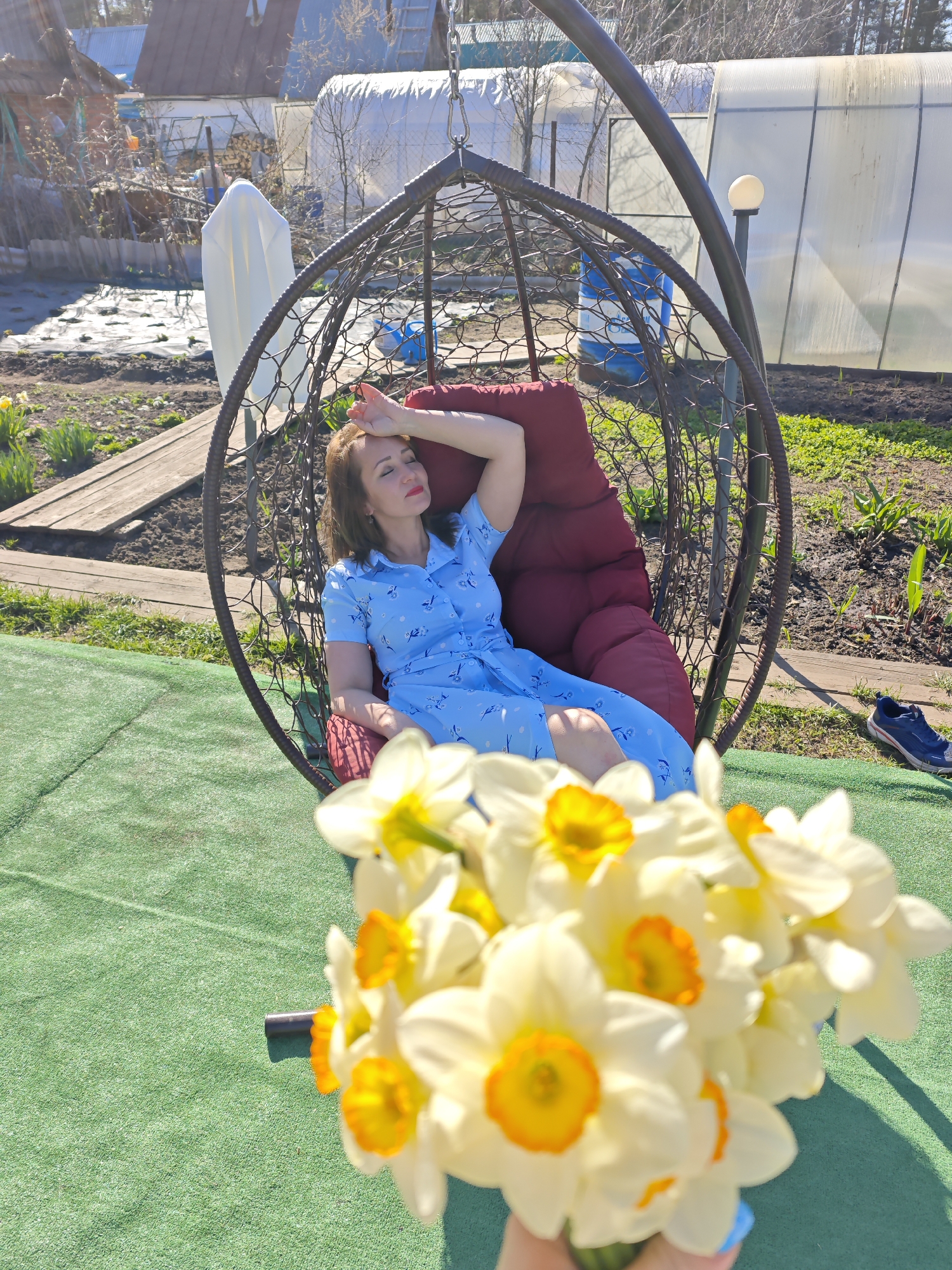 A woman in a blue dress reclines in a hanging chair. She has one hand on her forehead and her eyes are closed. In the foreground is a bouquet of daffodils. The background includes greenery, a greenhouse, and other structures. The picture was taken in Noviy, Russia.